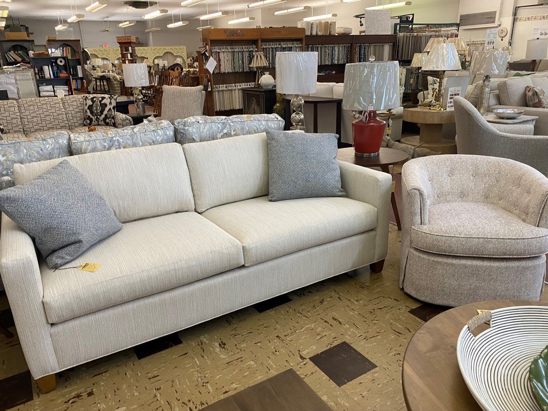 A cream-colored sofa with grey patterned throw pillows, a matching swivel chair, and lamps in a furniture showroom.