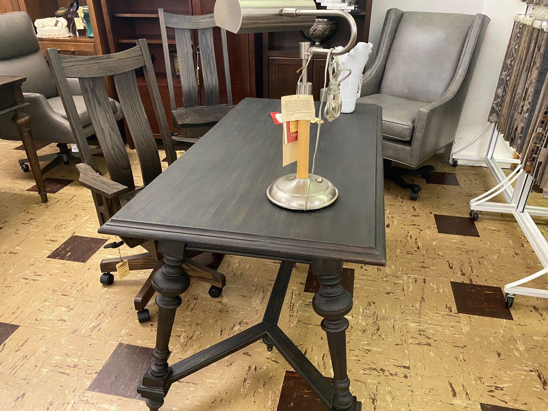 A dark wooden desk with ornate legs and a silver lamp sits in a room with office chairs and furniture displays.