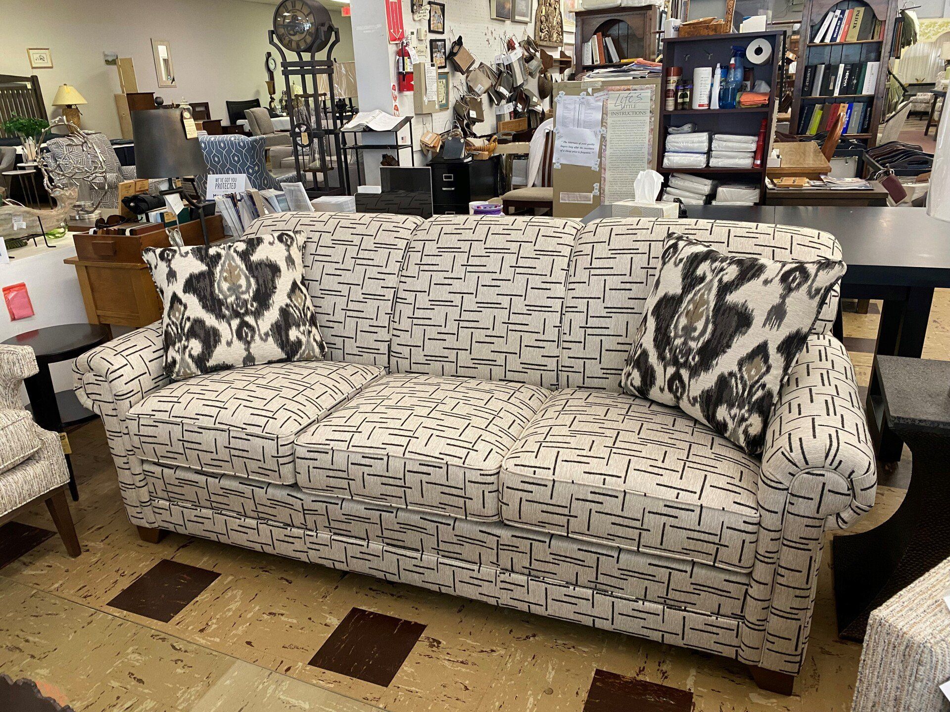 A three-seater sofa with an off-white and black geometric pattern, featuring two dark, patterned throw pillows in a store.