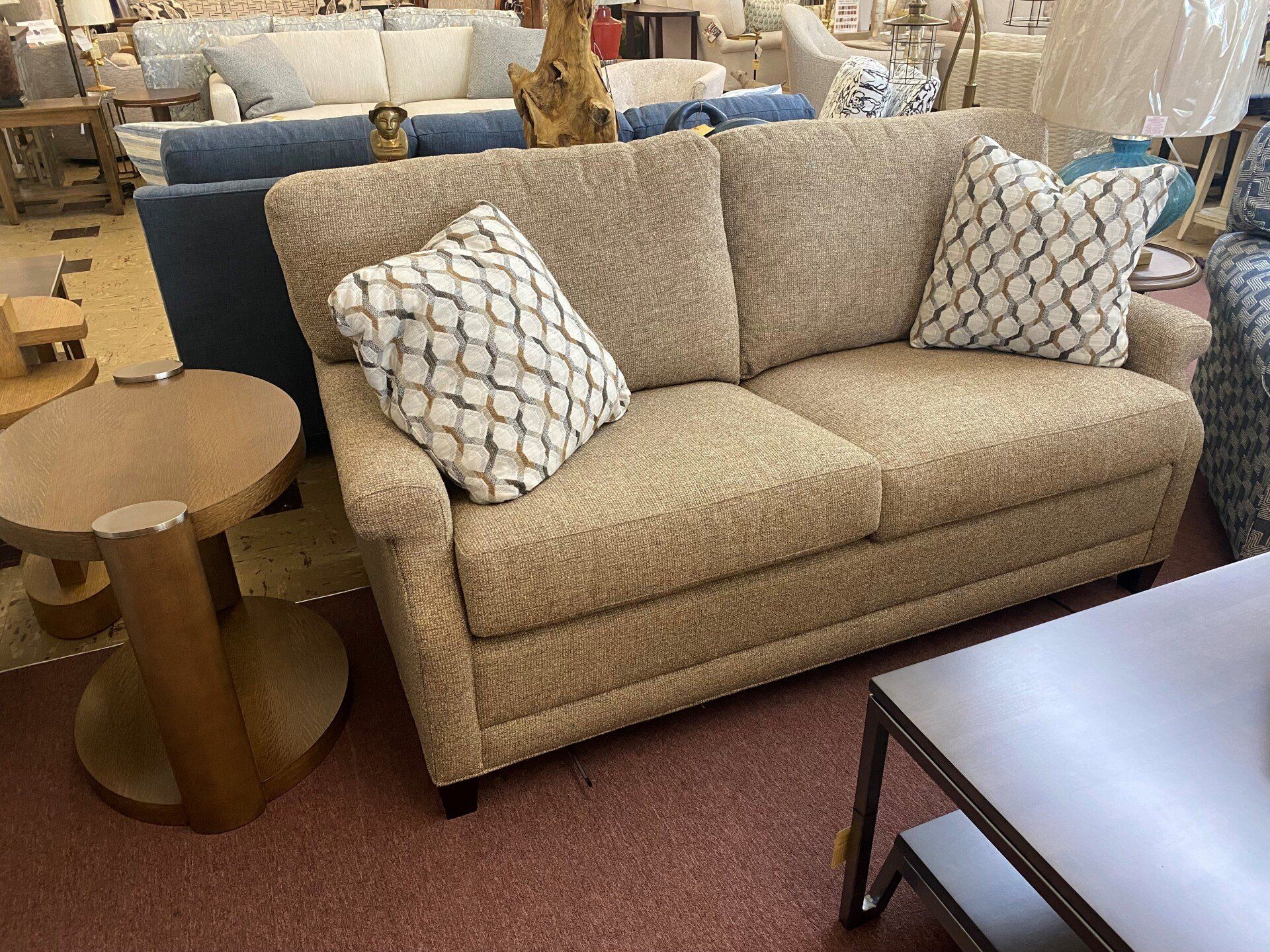 A textured tan couch with two patterned pillows sits in a furniture showroom next to a round wooden side table.