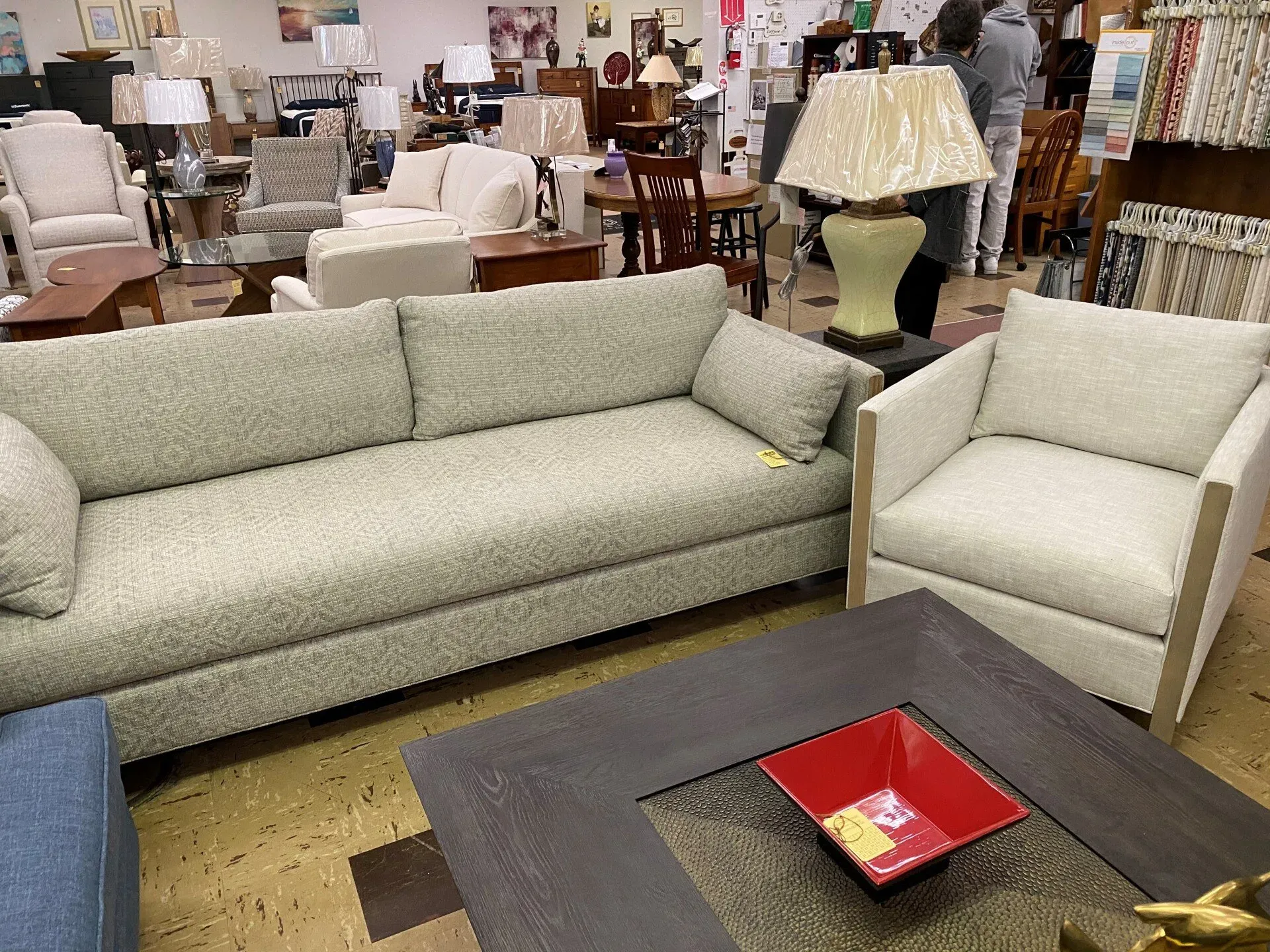 A showroom displays light-colored fabric couches and chairs arranged around a dark wooden coffee table with a red bowl.