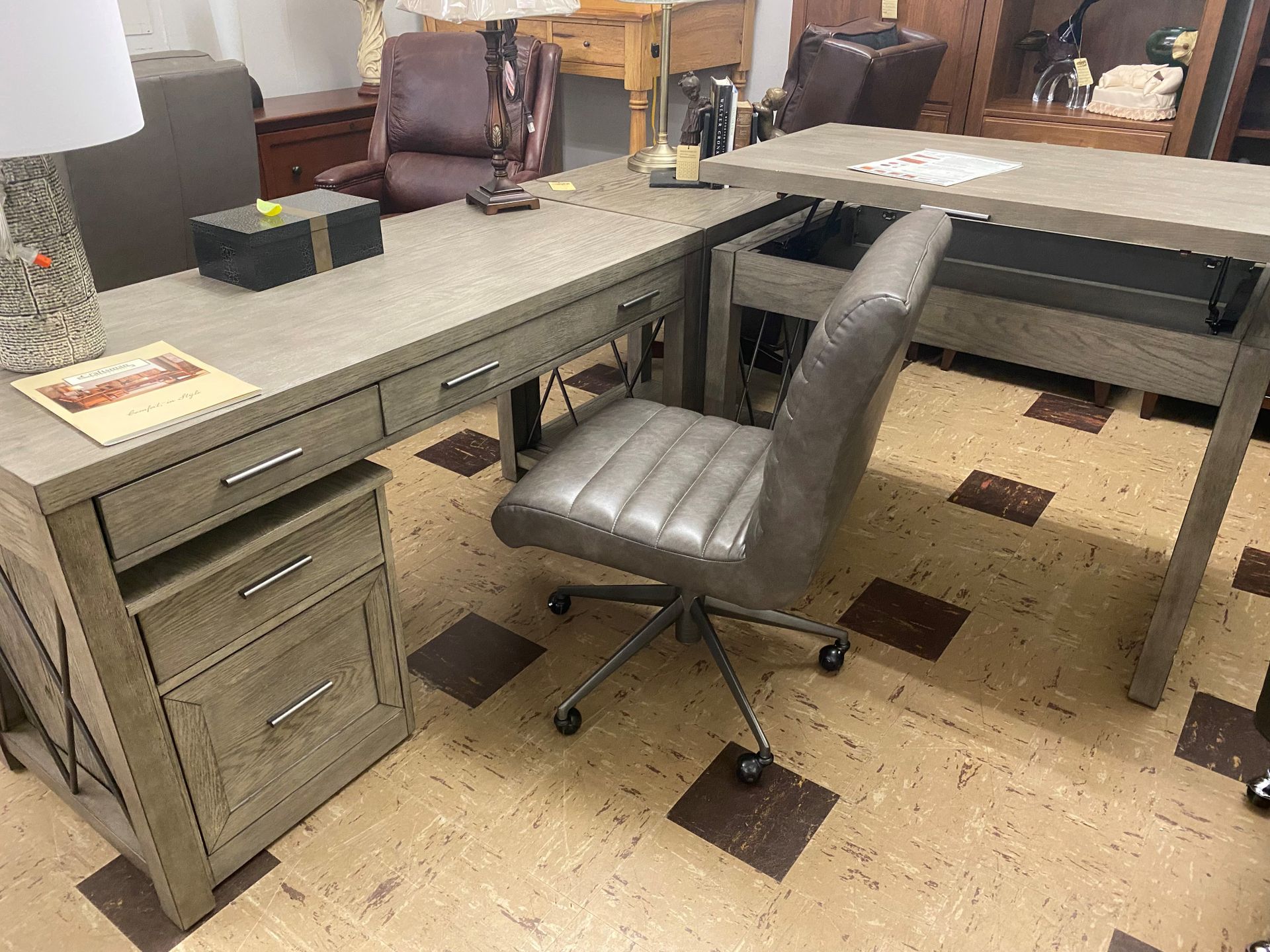 A grey wooden L-shaped desk with a matching grey office chair on wheels in a furniture store showroom.