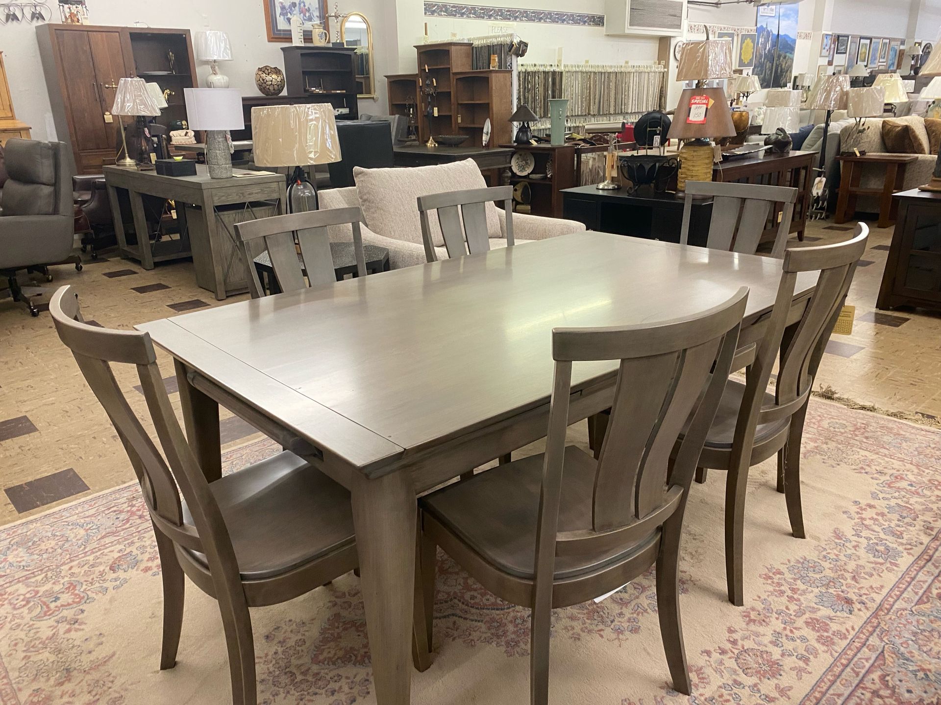 A grey wooden dining set with four matching chairs sits on an ornate rug inside a furniture store showroom.