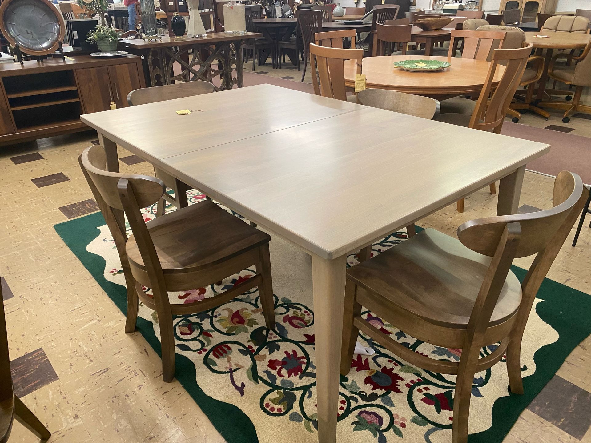 A light wood dining table with four matching chairs sits on a patterned rug in a furniture showroom.