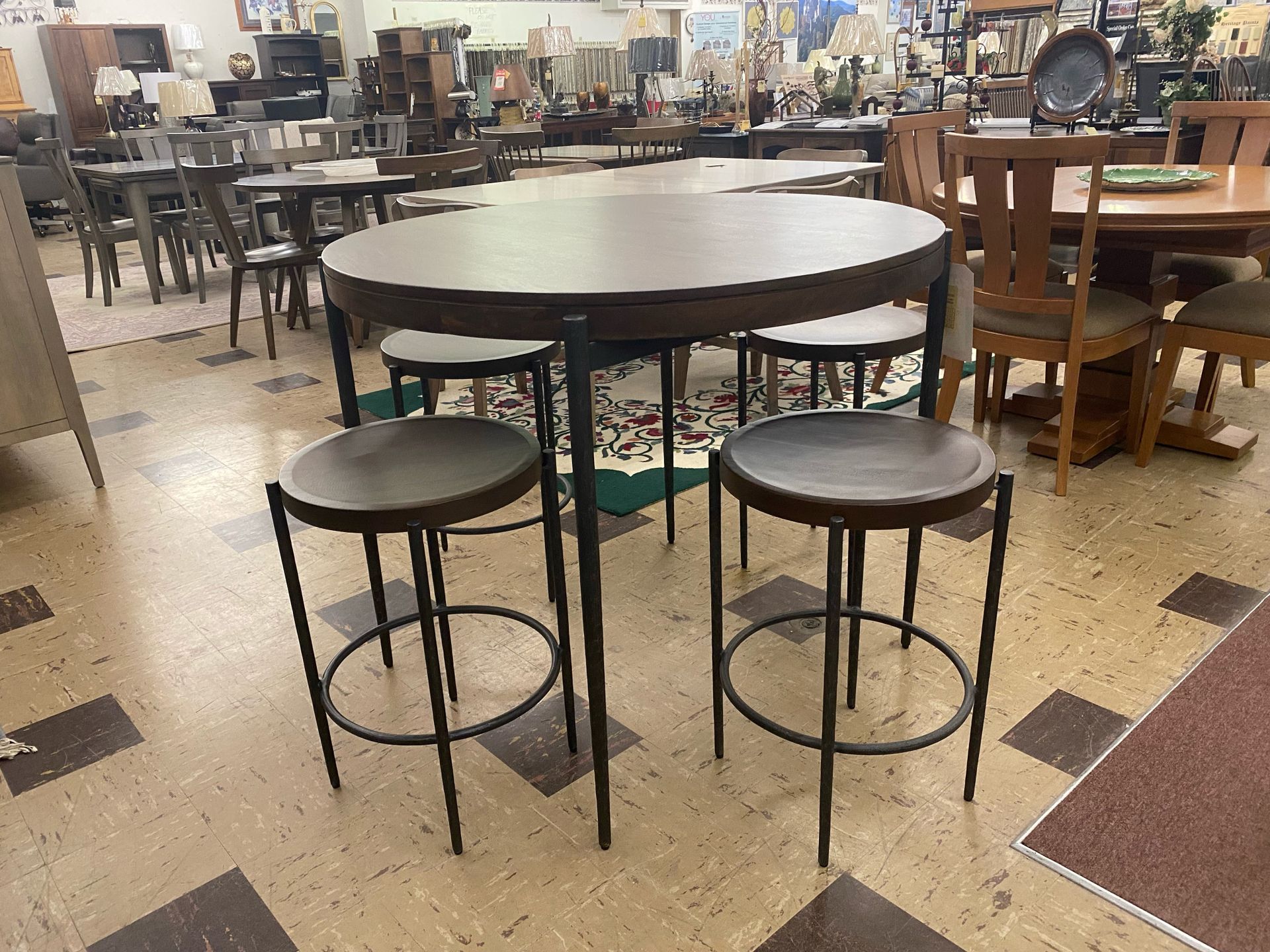 A small, circular, dark wood bistro table with four matching metal stools arranged in a retail furniture showroom.