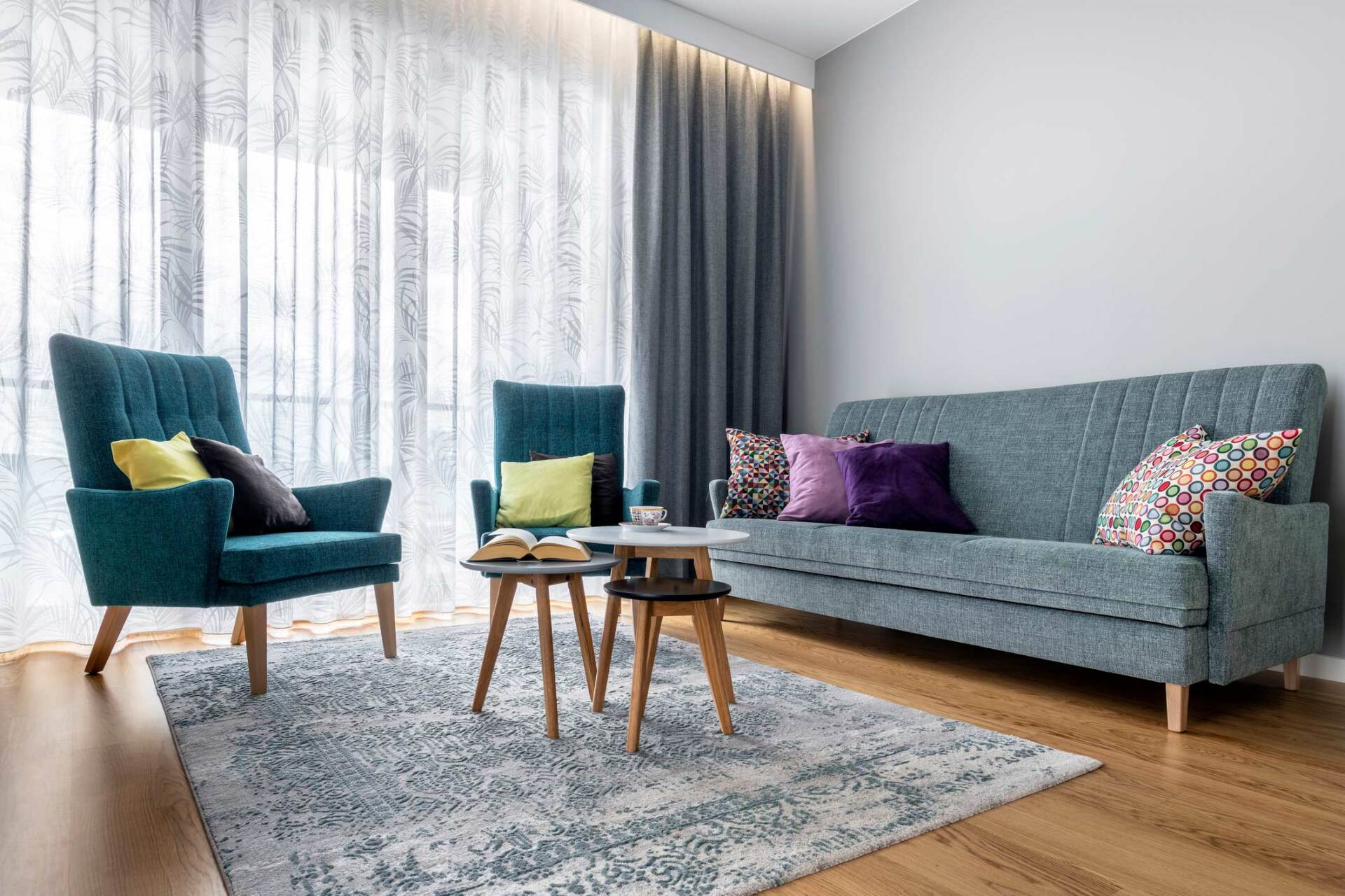 A cozy living room with a teal armchair, a smaller matching chair, and a teal sofa on a patterned rug, set near curtains.