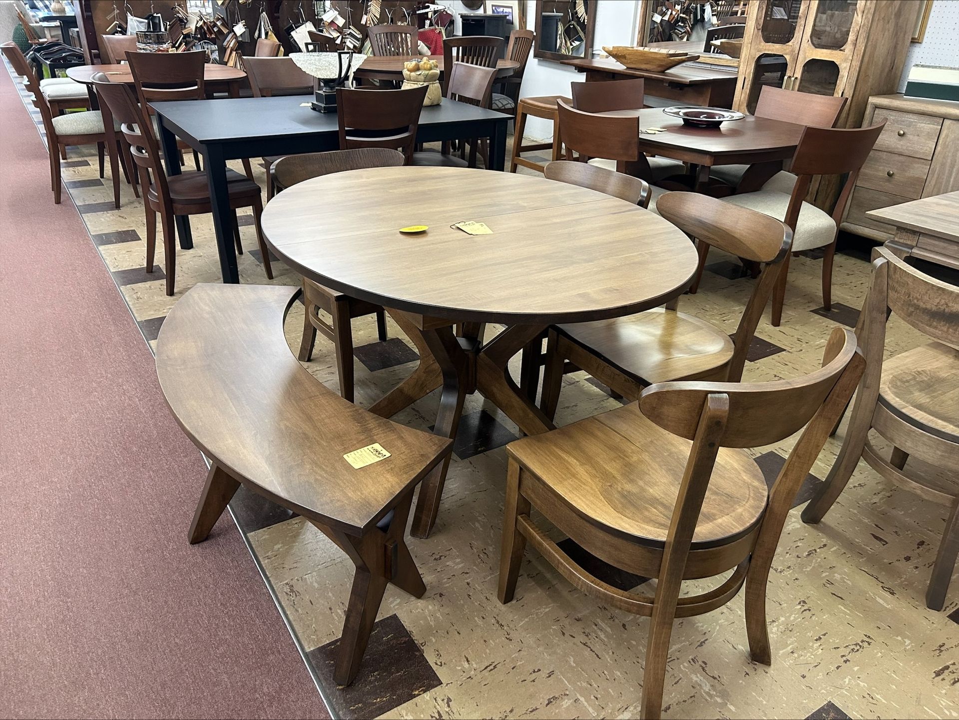A light-wood dining set featuring an oval table, two wooden chairs, and a curved bench in a furniture showroom.
