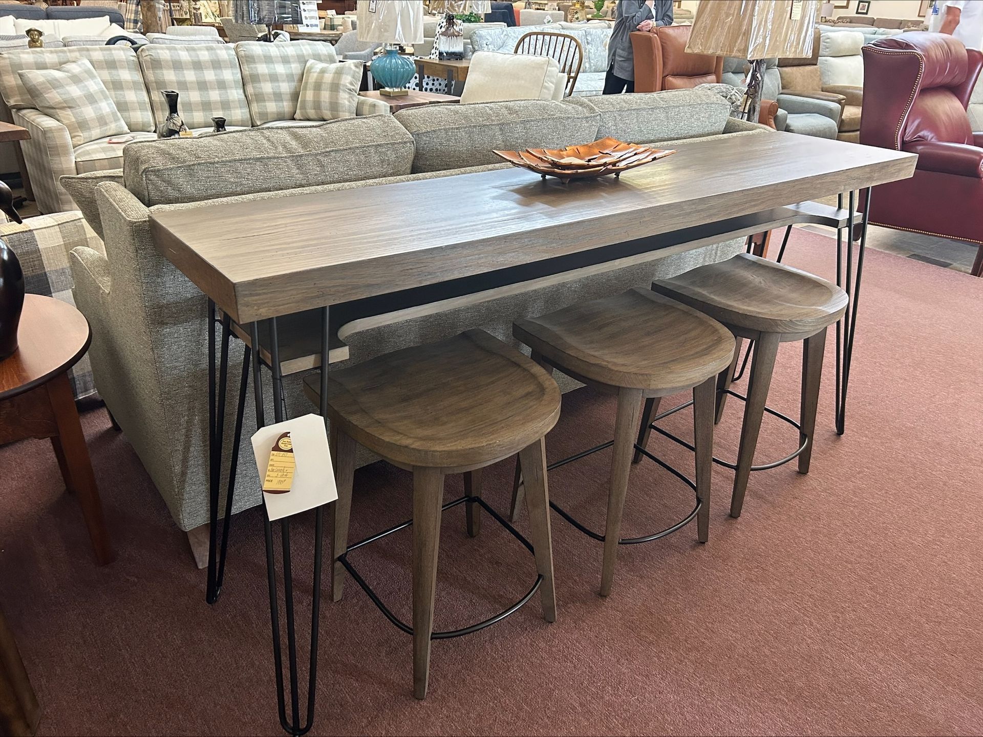 A tall, rectangular wooden sofa table with black metal hairpin legs and three matching barstools in a furniture store.