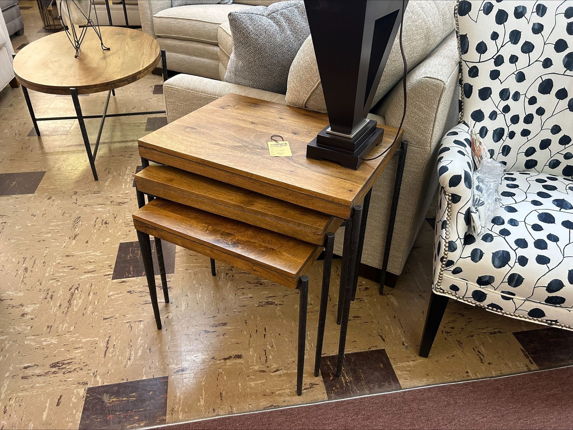 A nesting table set with wooden tops and dark metal legs, displayed in a furniture store next to a patterned armchair.