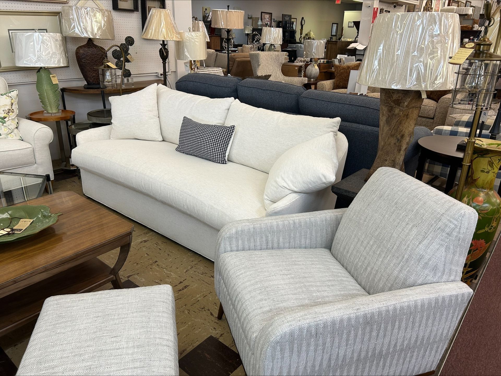 A showroom display features a white sofa, a light gray textured armchair with an ottoman, and several table lamps.