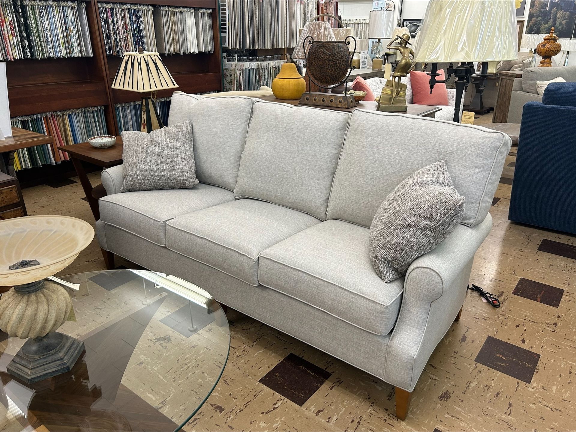 A light gray sofa with two patterned accent pillows sits in a showroom next to a glass-topped table and floor lamps.