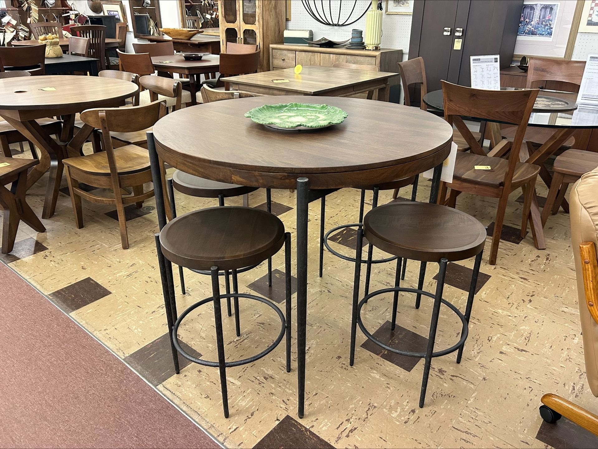 A round wooden pub-style table with a green plate, paired with two backless bar stools, in a furniture store showroom.