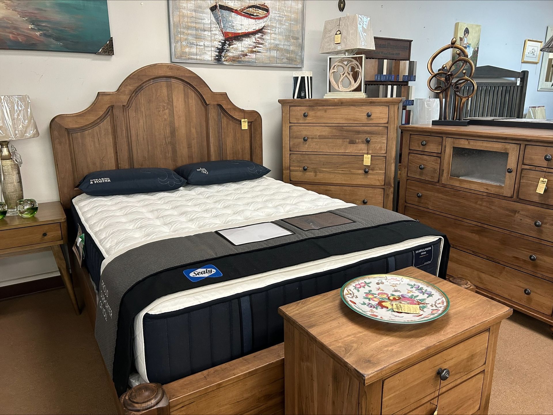 A bedroom display features a wood bed frame with a mattress, two matching nightstands, and a tall chest of drawers.