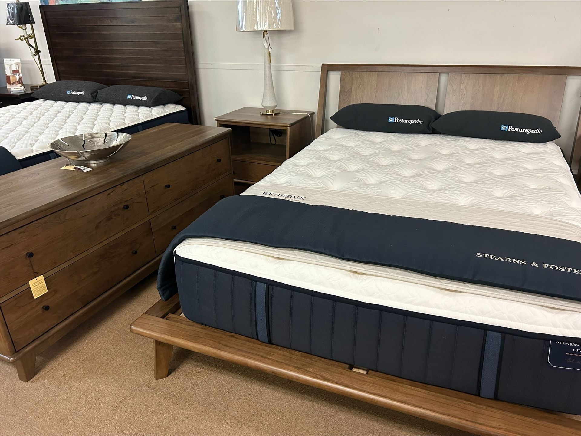 A showroom display features two beds with dark wood frames and white mattresses, separated by a wooden nightstand.