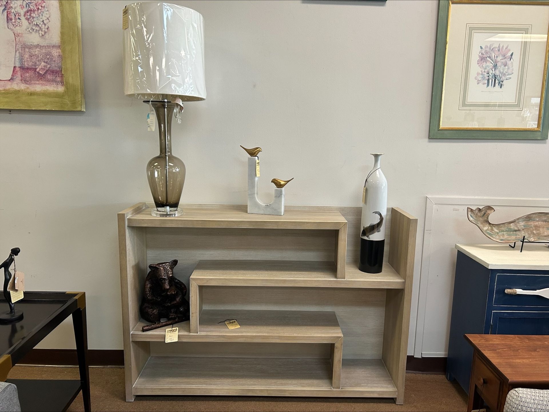 A light-wood tiered shelf sits against a wall with a lamp, bird figurines, and a decorative vase on top.