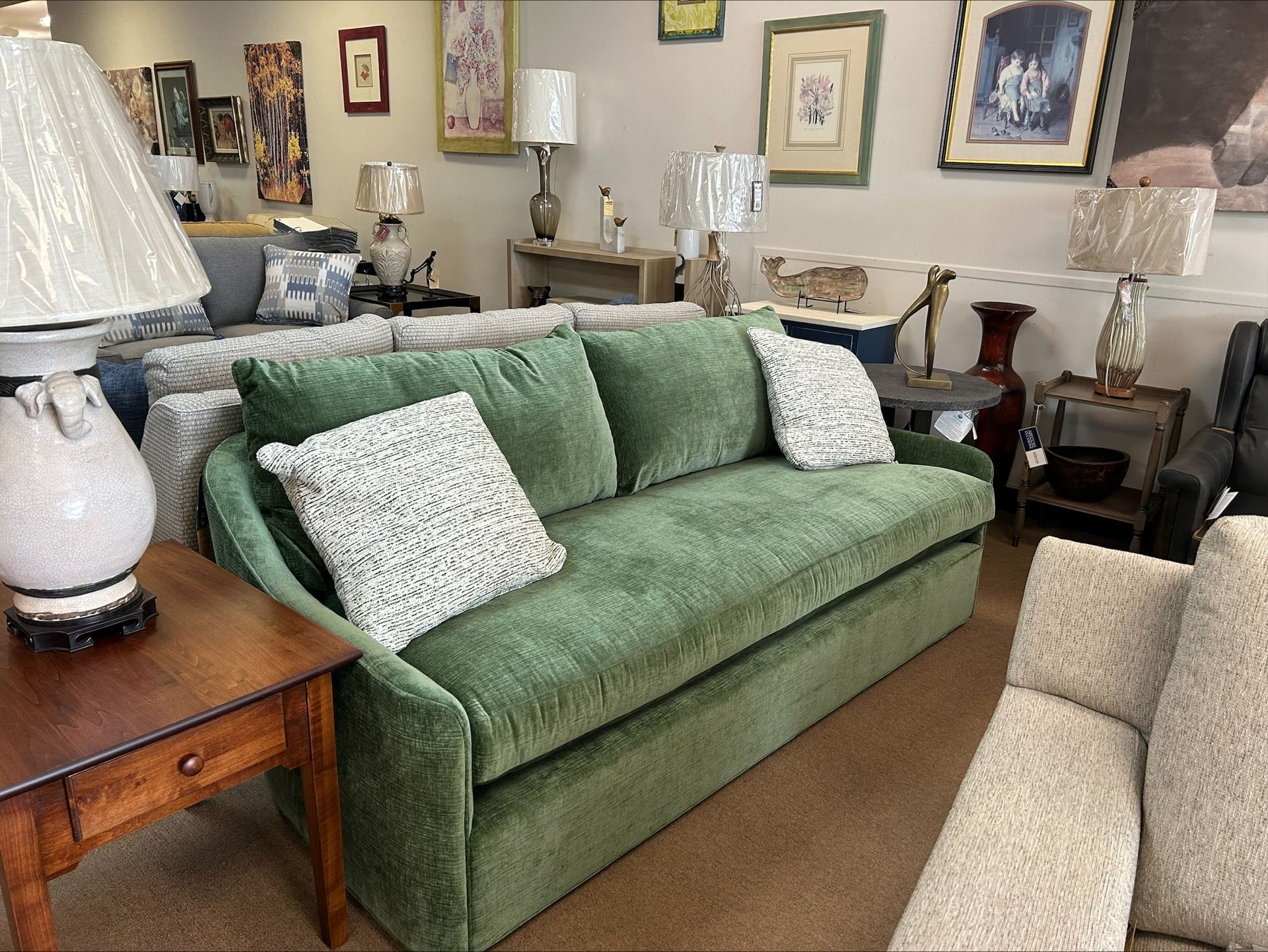 A furniture showroom featuring a prominent sage green couch with patterned pillows, lamps, side tables, and wall art.