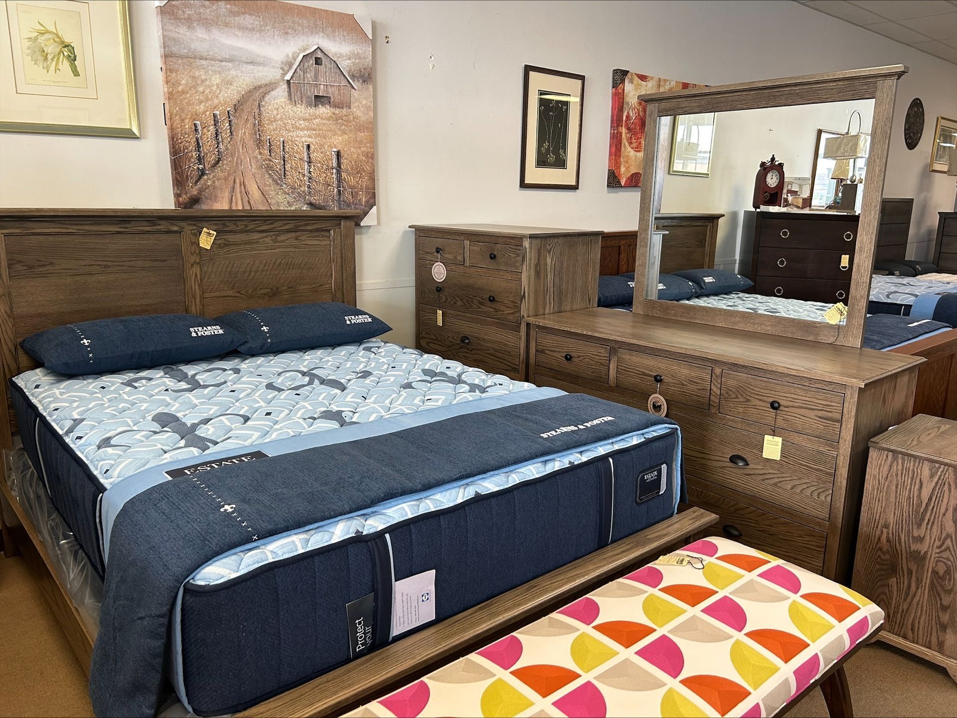 A bedroom set with a wooden bed, dresser with mirror, and a colorful patterned bench in a showroom.