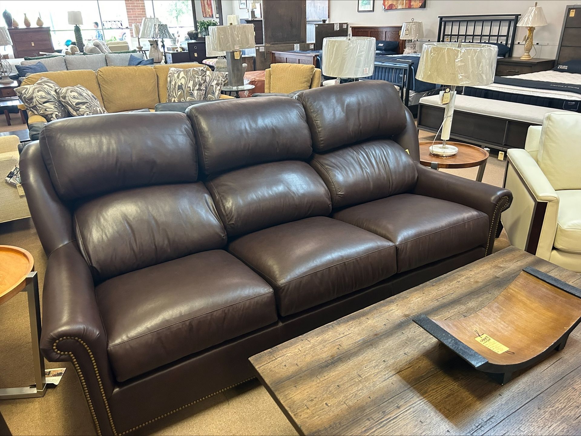 A three-cushion, dark brown leather sofa with nailhead trim sits in a furniture showroom next to a coffee table.
