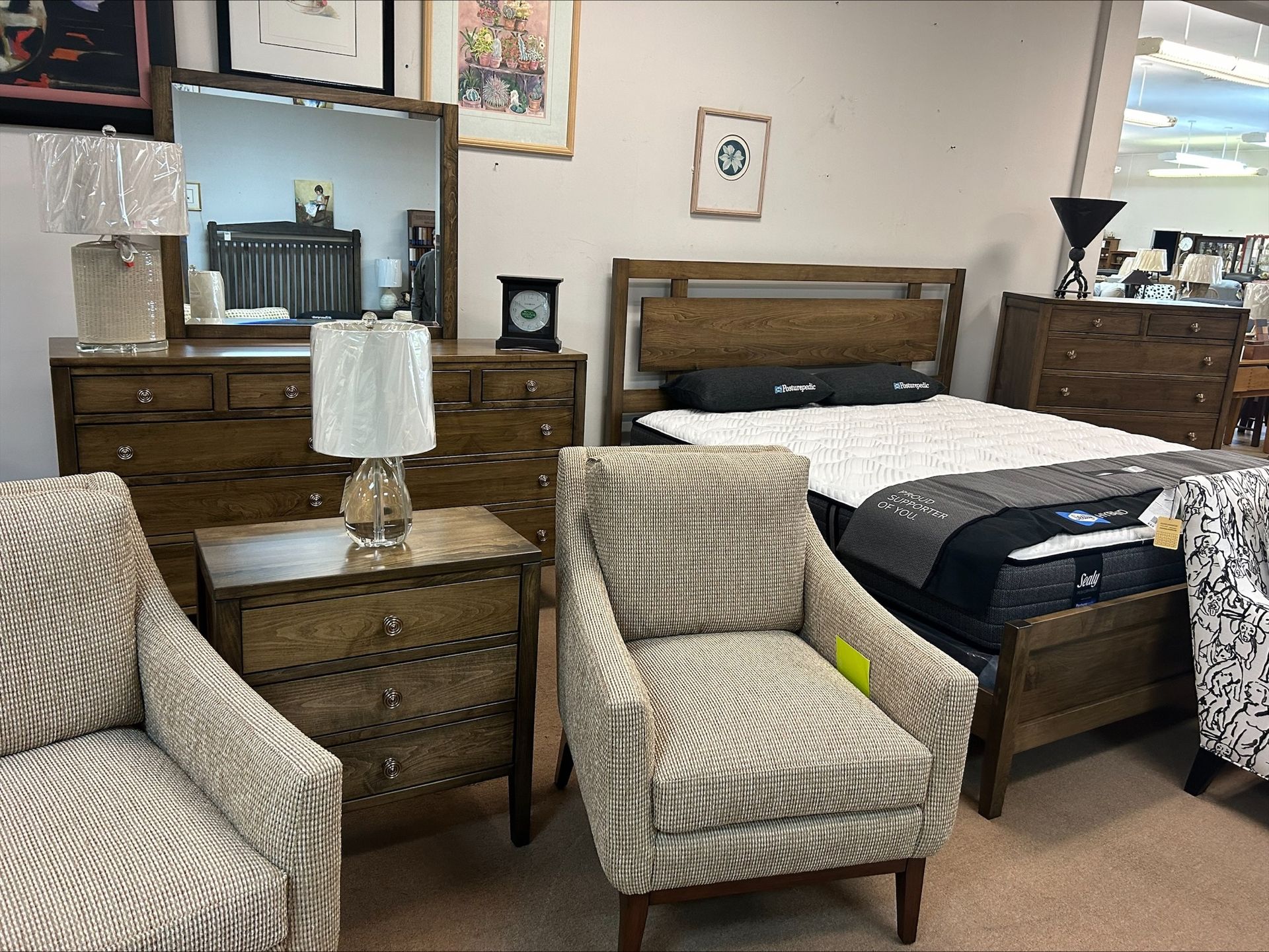 A furniture store showroom displays a wooden bedroom set, including a bed, dresser with mirror, nightstand, and two chairs.