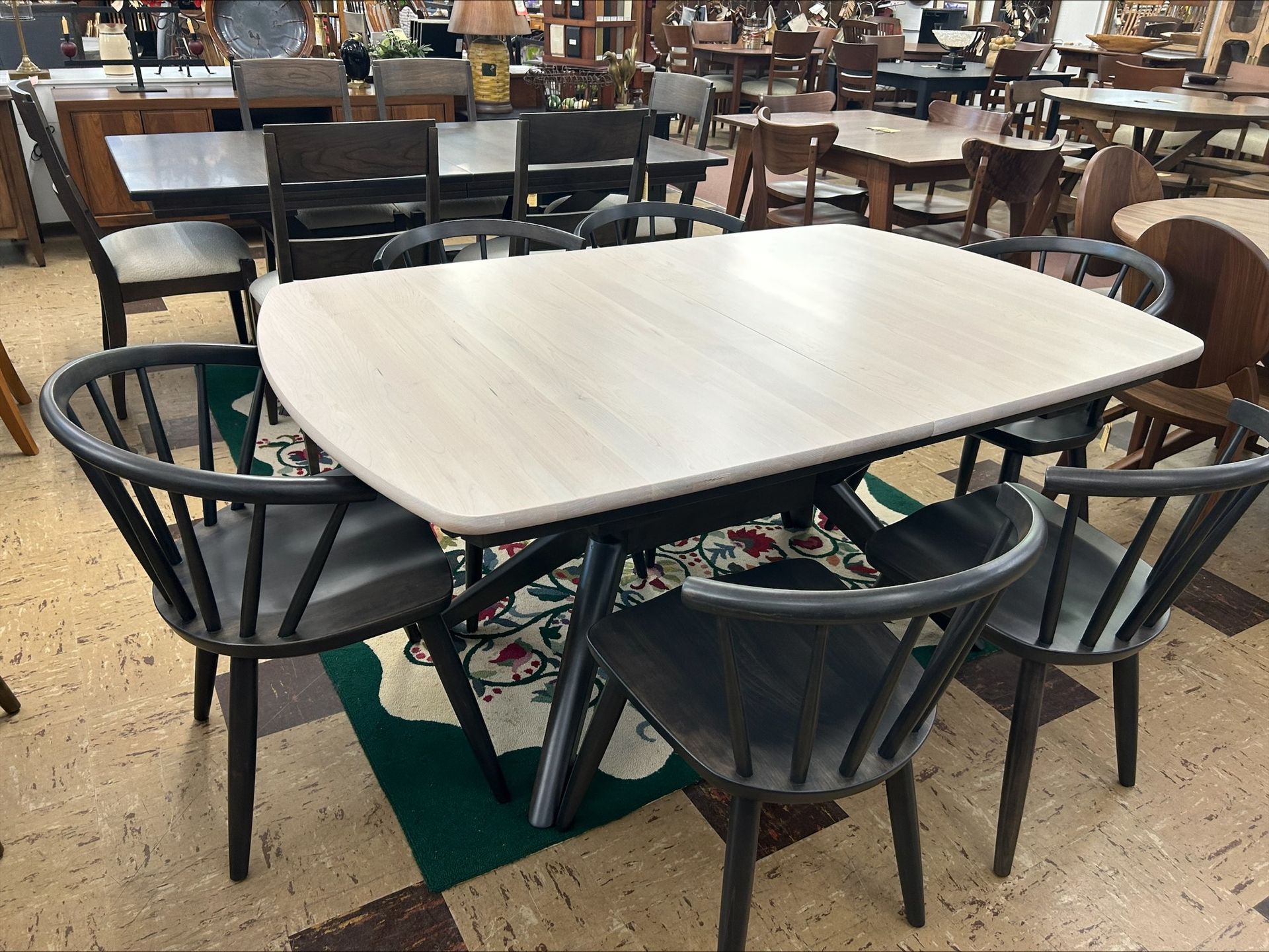 A light wood rectangular dining table with dark spindles chairs arranged on a patterned rug in a furniture showroom.
