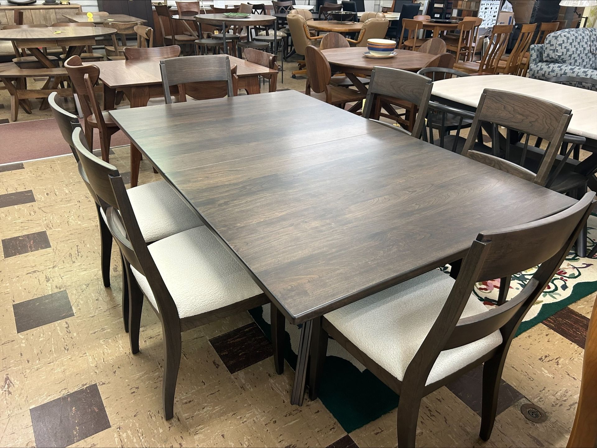 A rectangular dark wood dining table with chairs featuring beige upholstered seats, set in a showroom furniture store.