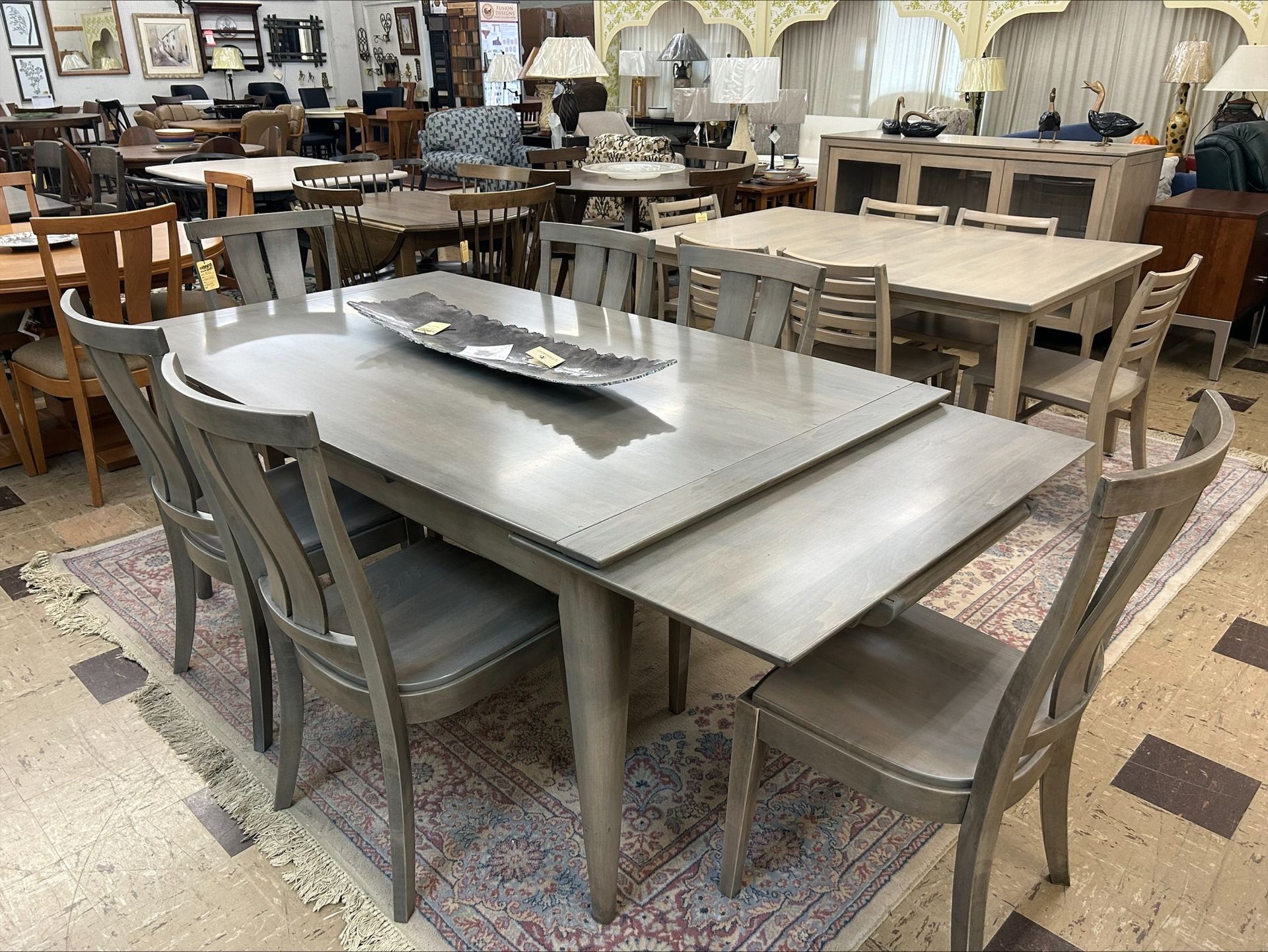 A grey dining table with chairs in a furniture showroom, featuring a center decorative tray on a patterned area rug.