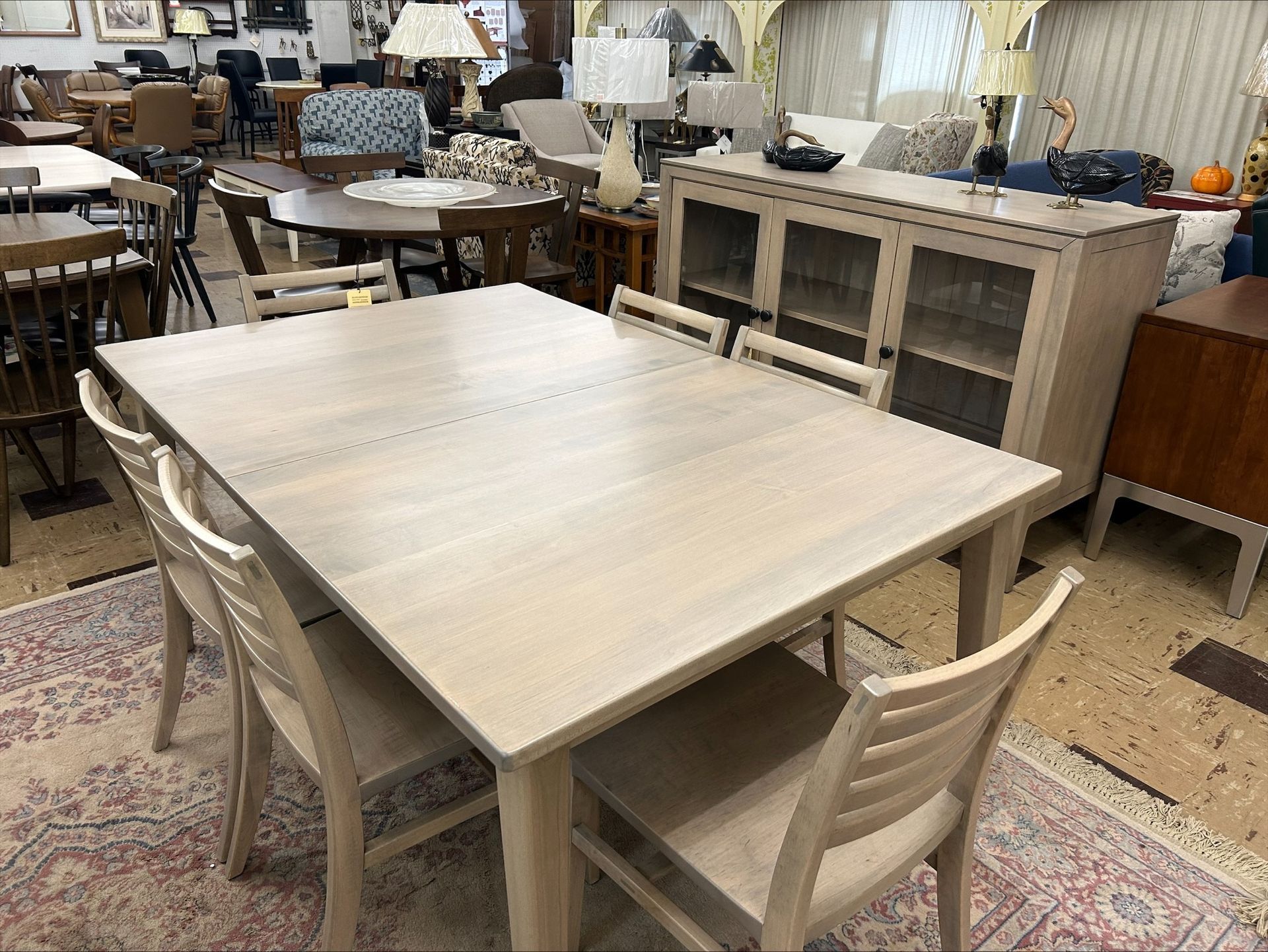 A light-wood dining set with four slat-back chairs sits on a patterned area rug in a furniture showroom.