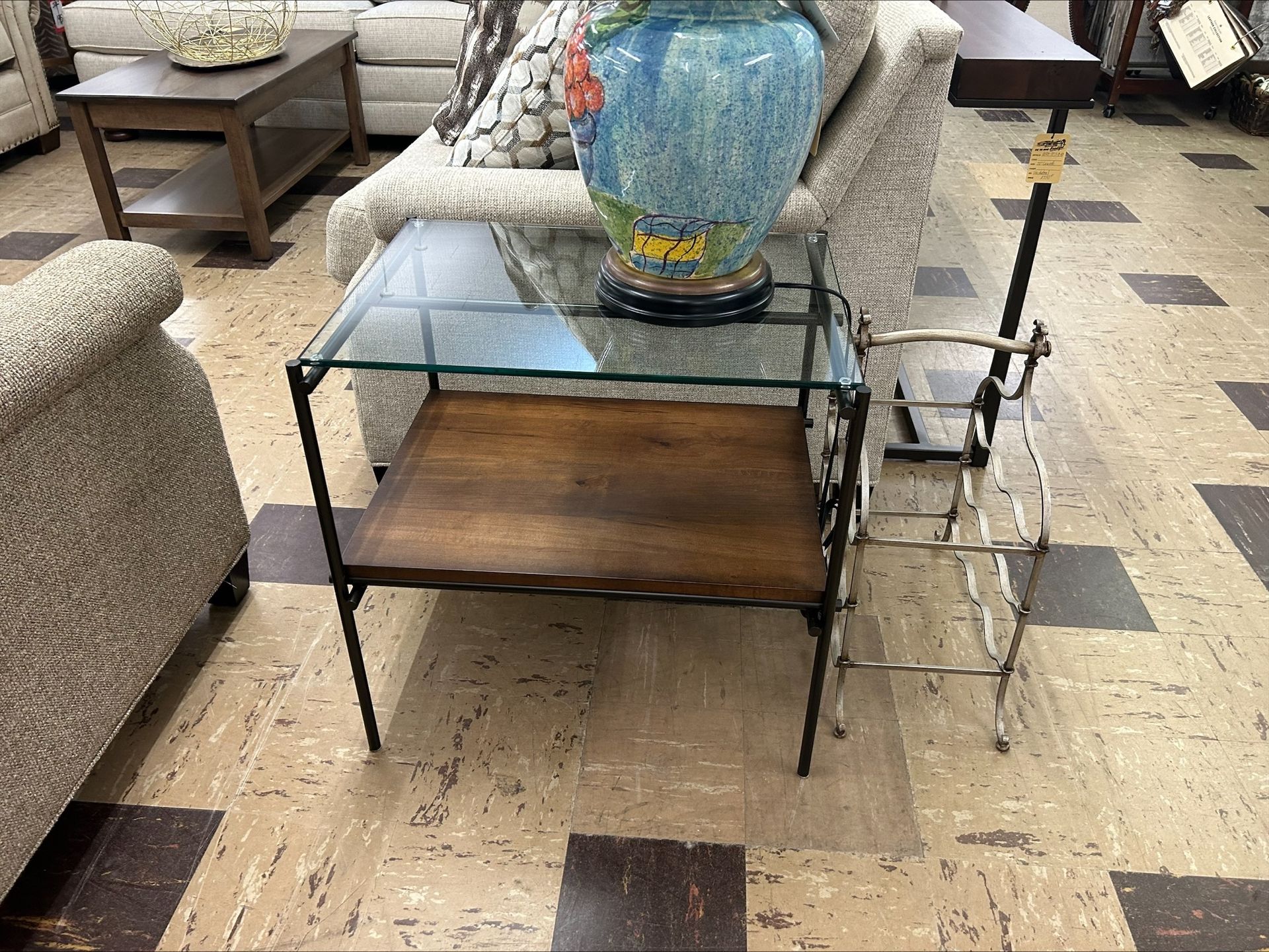 A square end table with a glass top and wooden lower shelf stands on a tile floor in a furniture store.