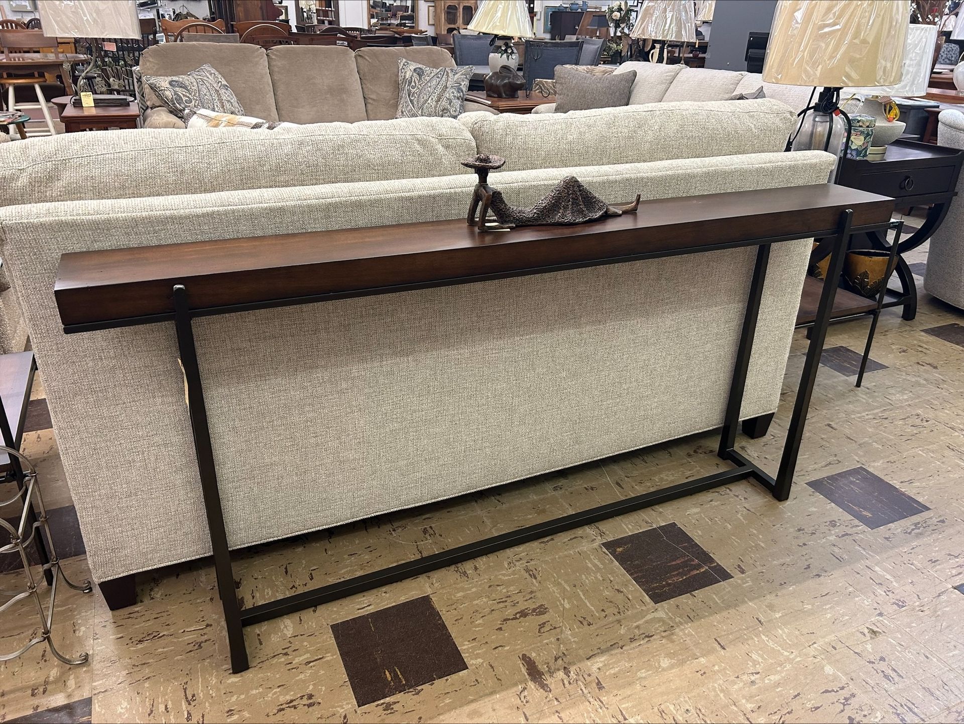 A slim, dark wood sofa table with a metal frame stands in front of a neutral-colored fabric sofa in a furniture store.
