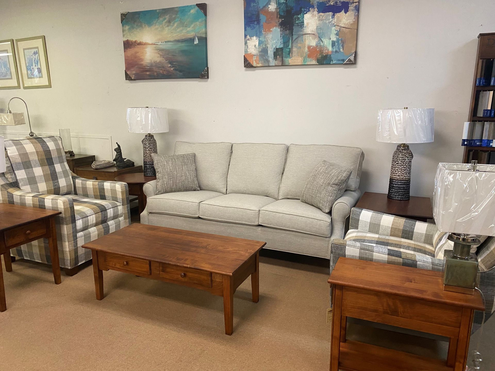 A beige sofa and two plaid armchairs in a showroom with wooden coffee and end tables, lamps, and wall art.