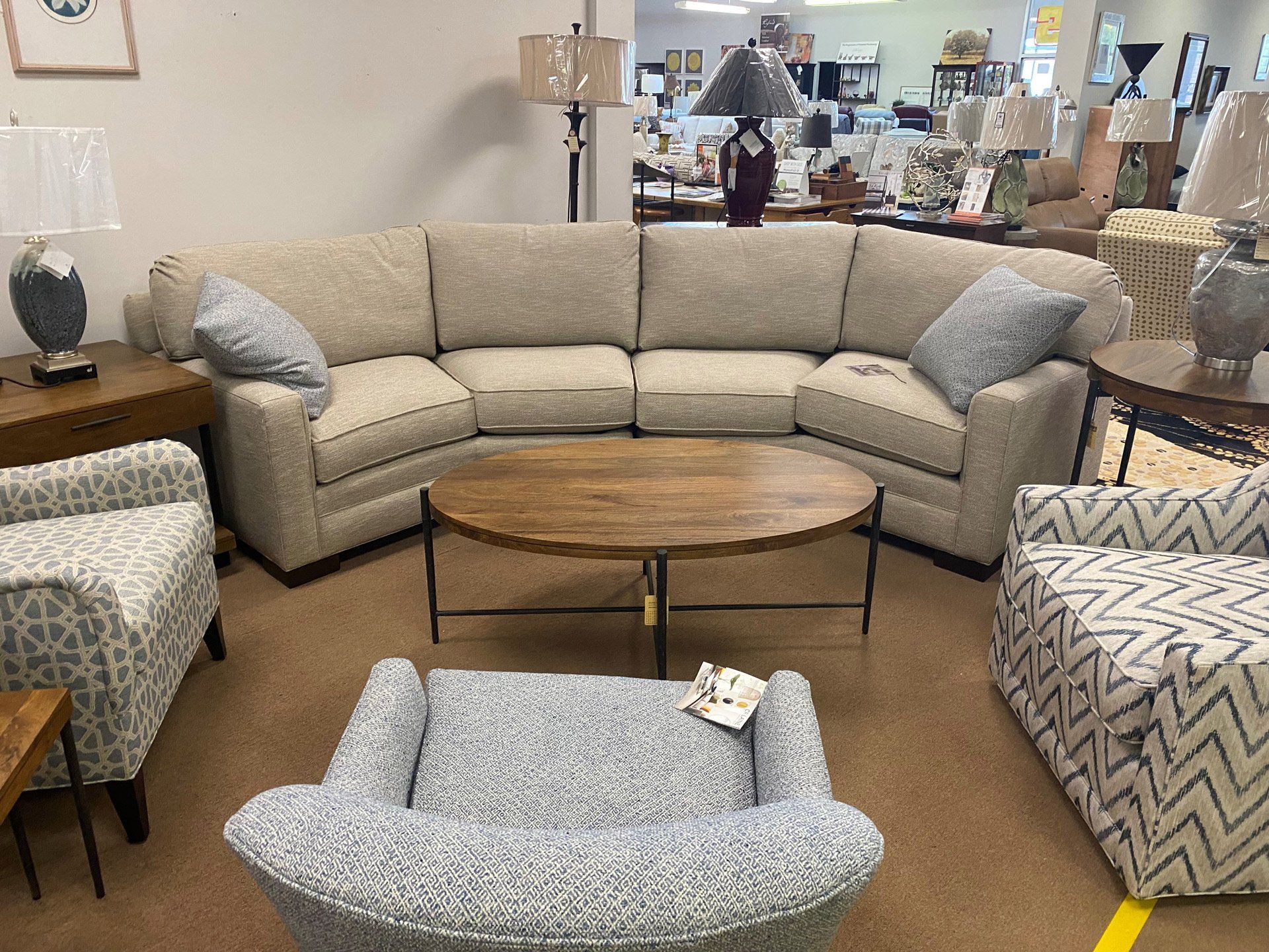 A showroom display featuring a curved, light-tan sectional sofa, three patterned armchairs, and a round wooden coffee table.