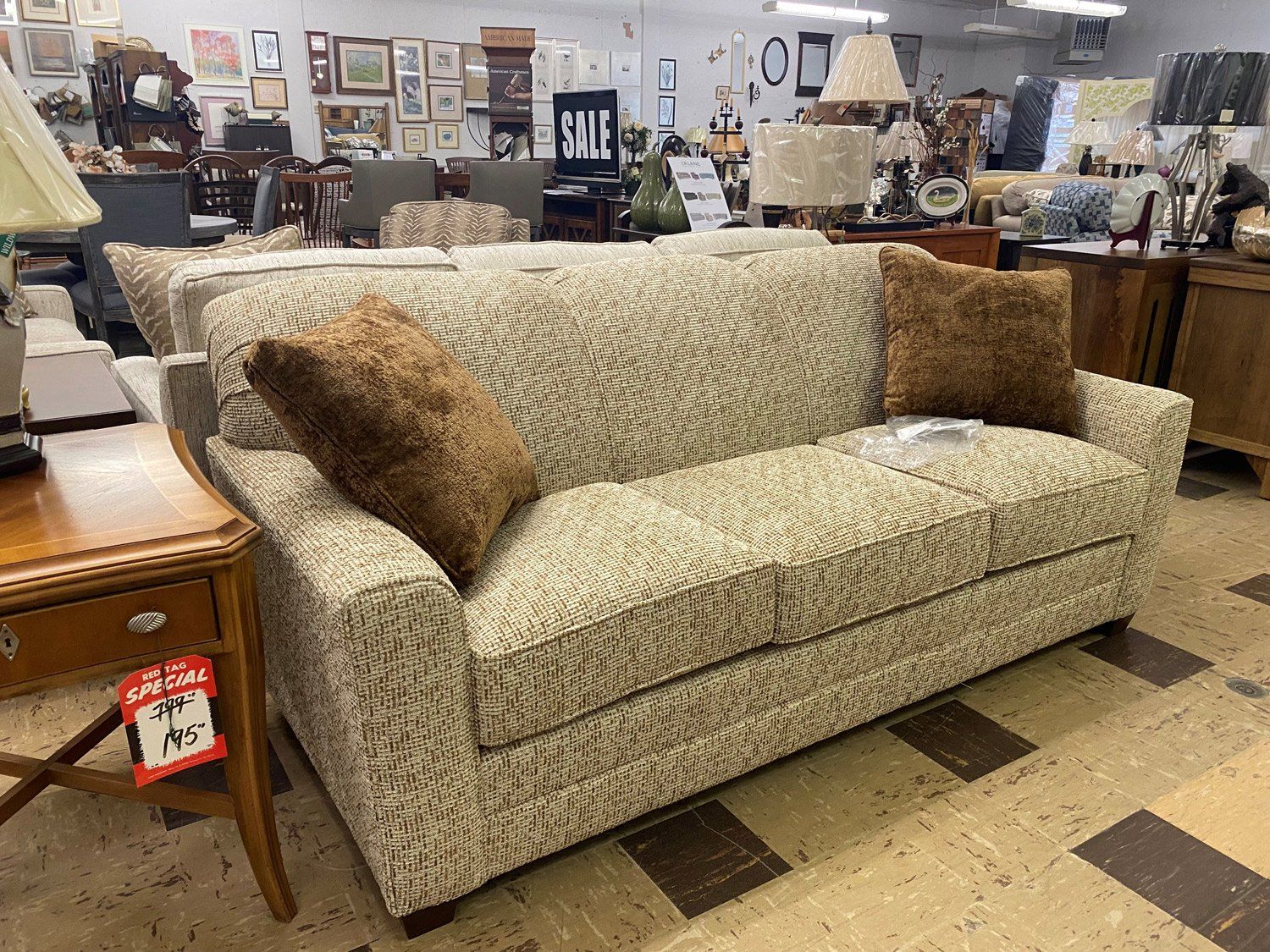 A patterned beige and brown sofa with two brown throw pillows sits on a tiled floor in a furniture store.