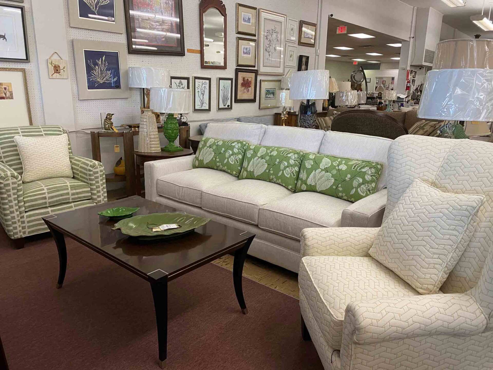 A furniture showroom featuring a light-colored sofa with green patterned pillows, two armchairs, and a dark wood coffee table.