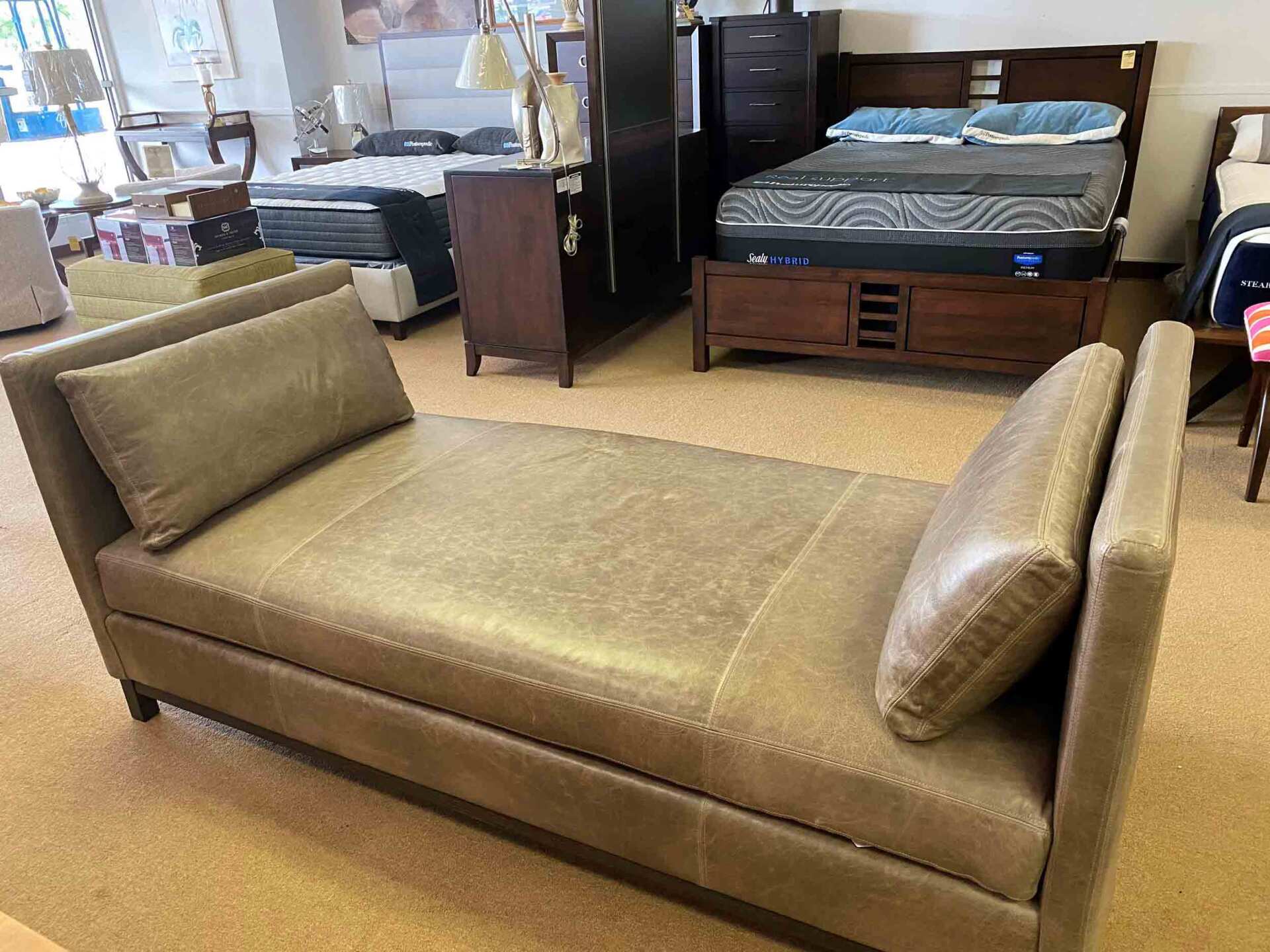 A beige, tufted leather daybed with two bolster pillows, displayed in a furniture showroom with beds in the background.