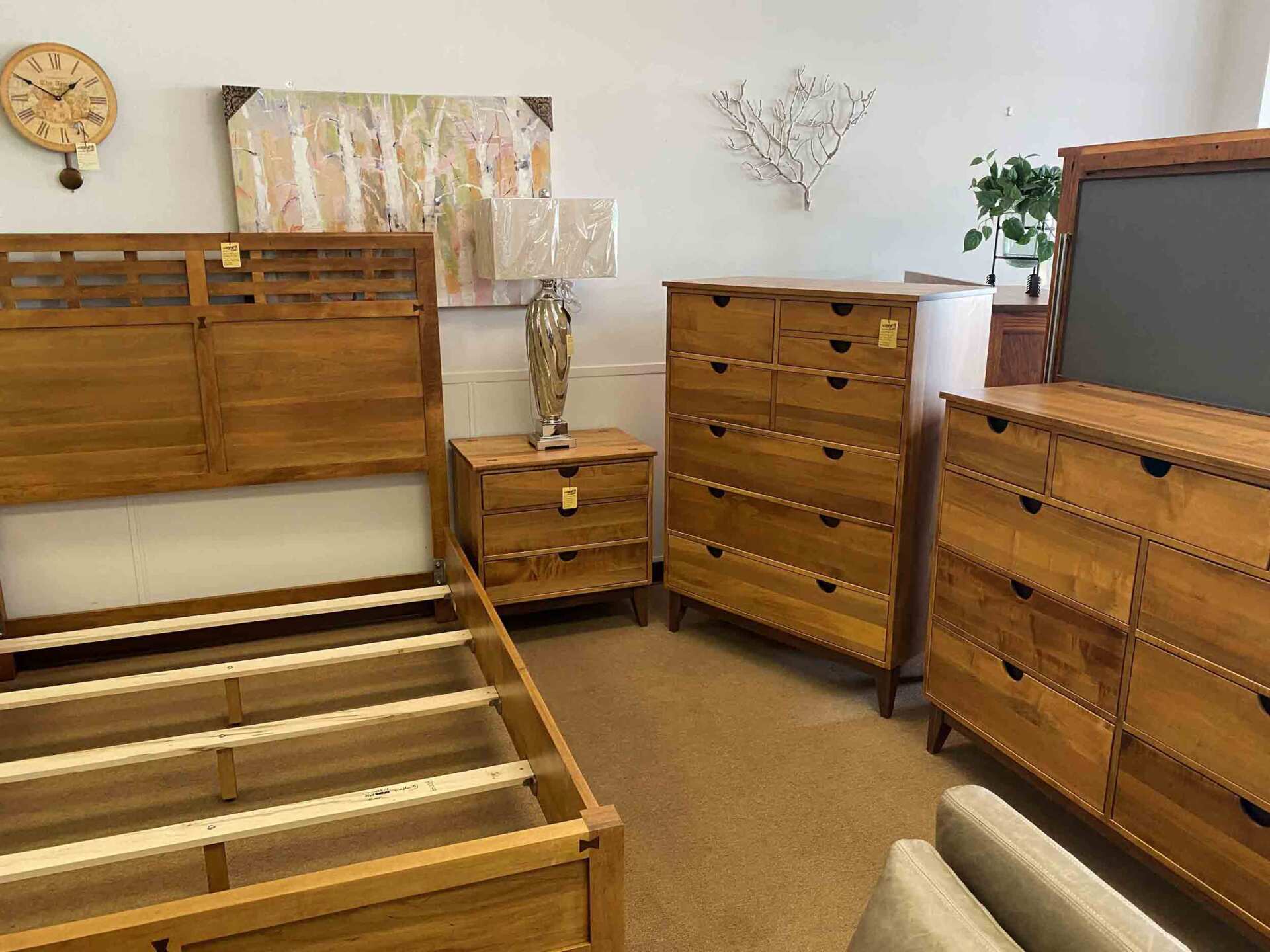 A showroom display featuring a wooden bed frame, nightstand, tall chest, and dresser with a mirror against a beige wall.