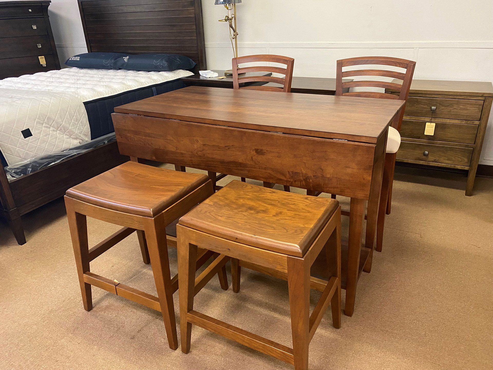 A wooden drop-leaf table with two stools and two chairs in a store, situated next to a bed and a dresser.
