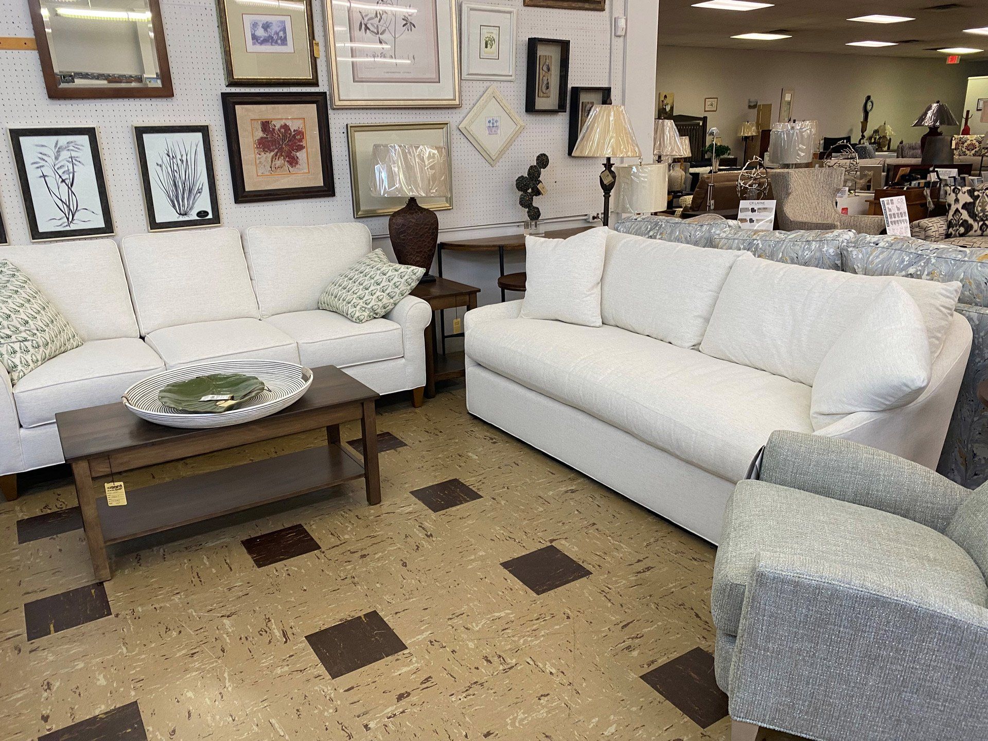 Two white sofas arranged in a showroom with a wooden coffee table and a gray armchair in the foreground.