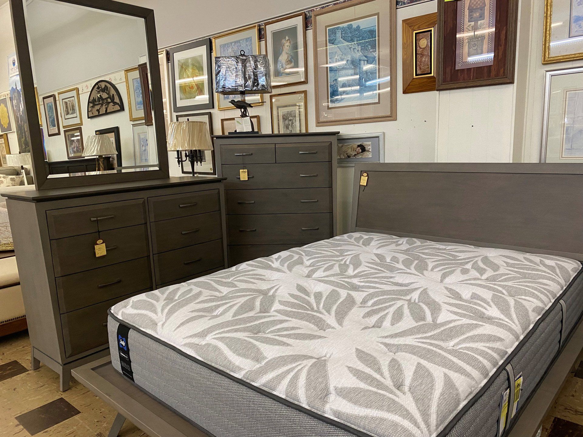 A display of grey bedroom furniture, including a bed with a patterned mattress, a dresser with a mirror, and a chest.