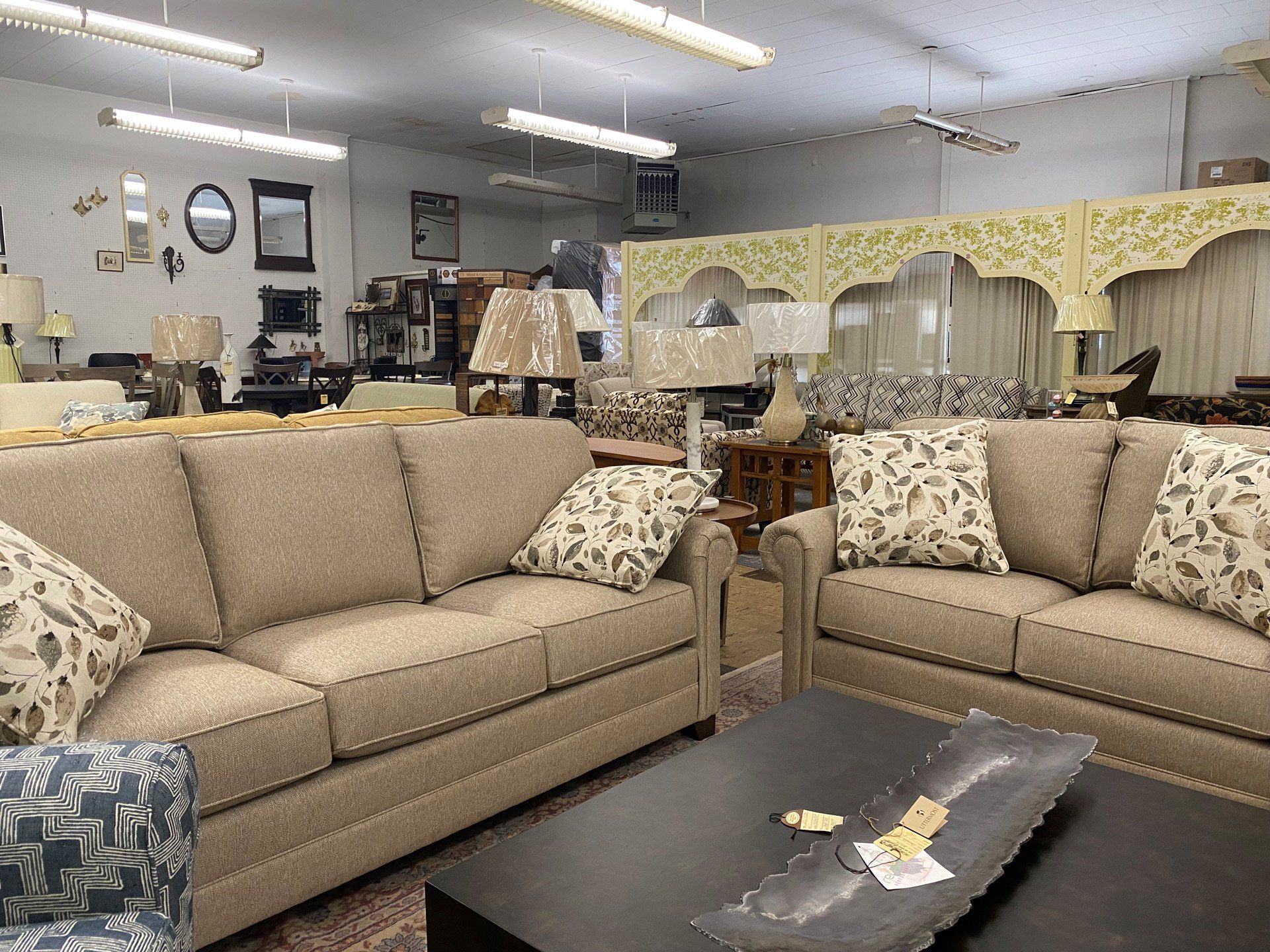 A furniture showroom featuring two neutral-toned fabric sofas, decorative throw pillows, and several lamps.