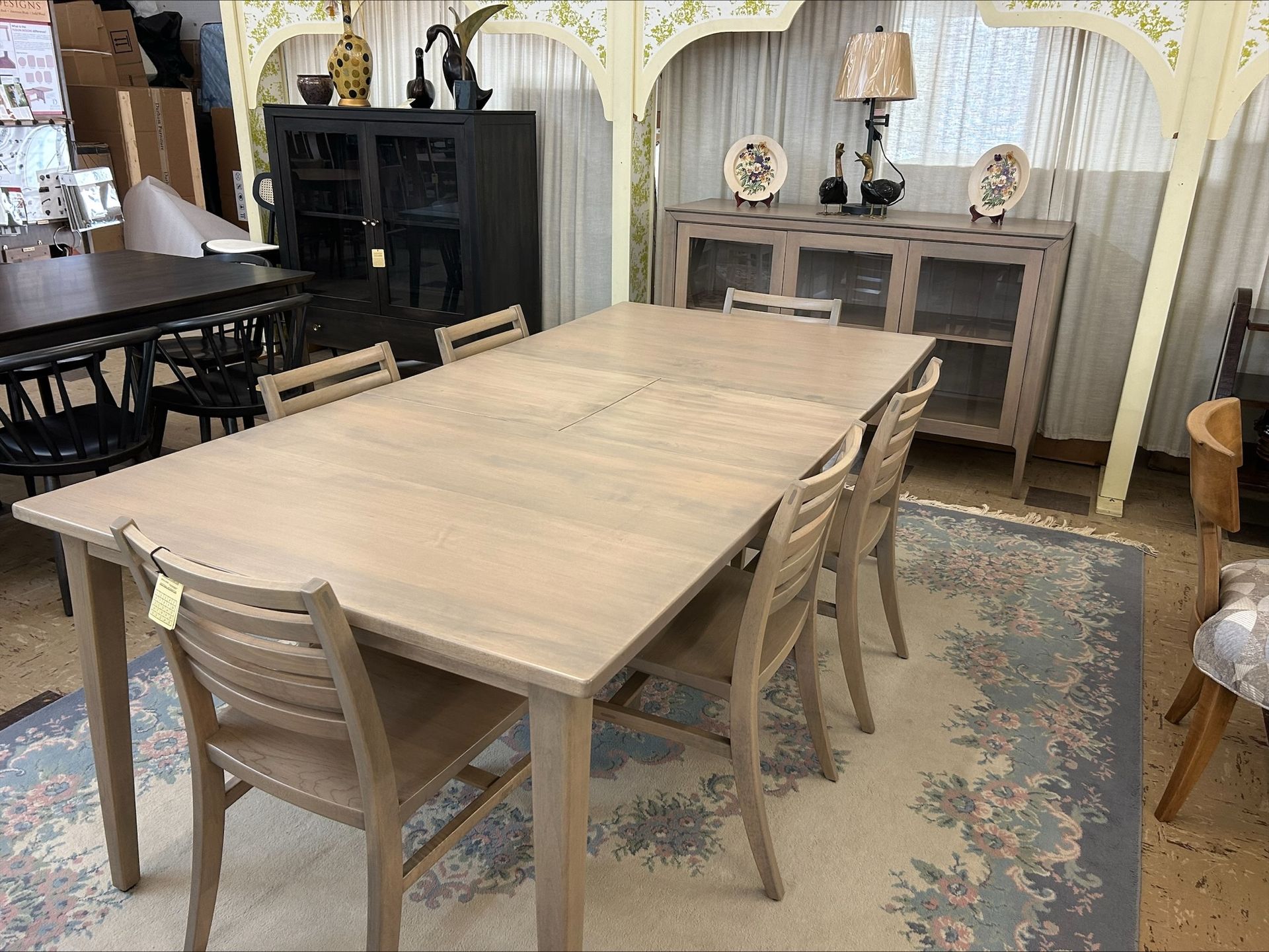 Light wood dining table and chairs in a decorated furniture showroom with rugs and cabinets.