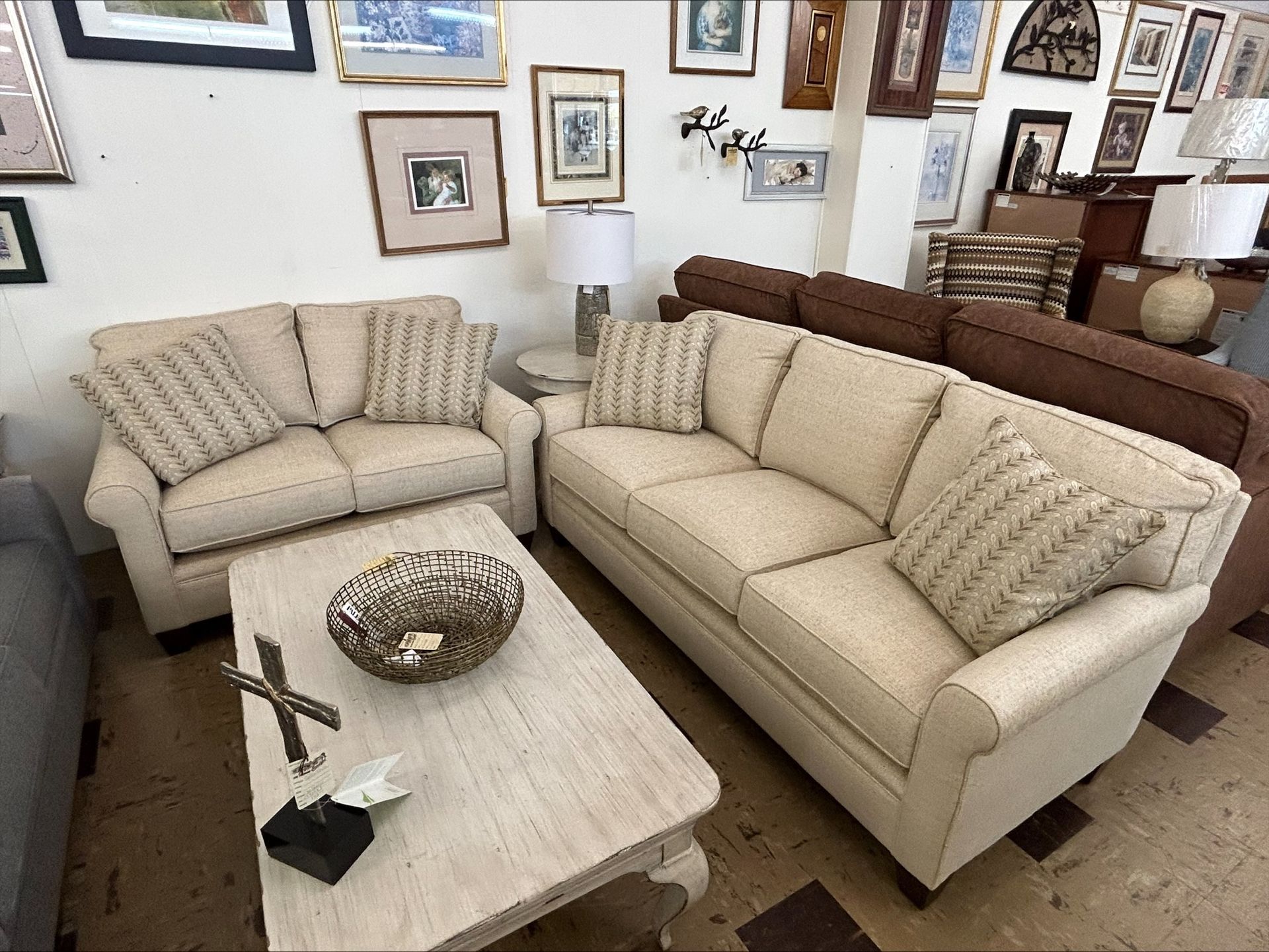A beige sofa and loveseat set arranged with a light wood coffee table in a furniture showroom with wall-mounted decor.