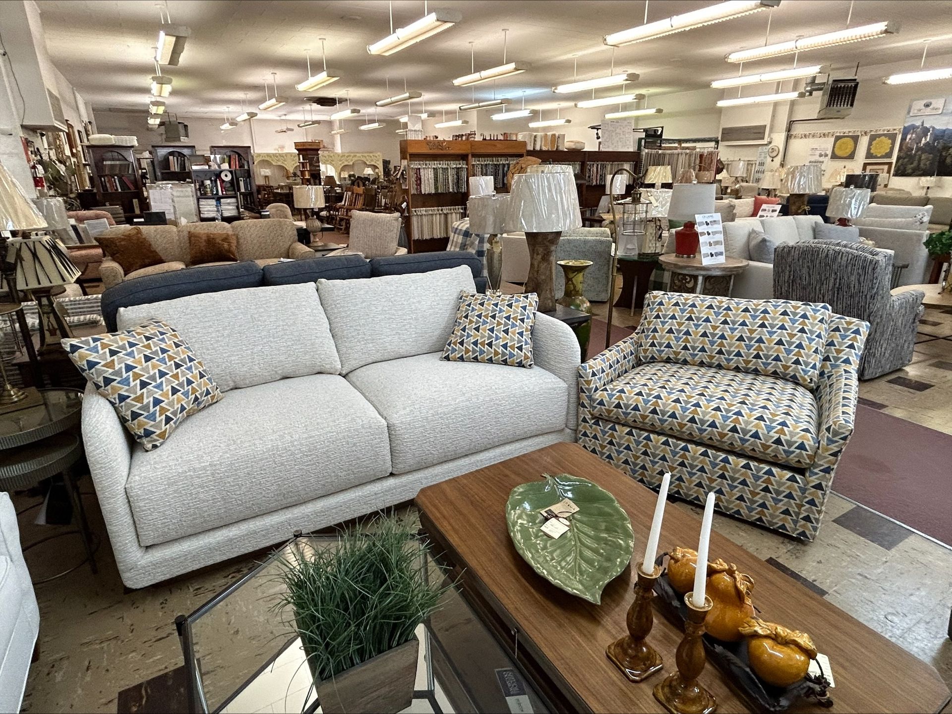 A showroom display with a light gray sofa and a patterned armchair, featuring a coffee table with decor in the foreground.