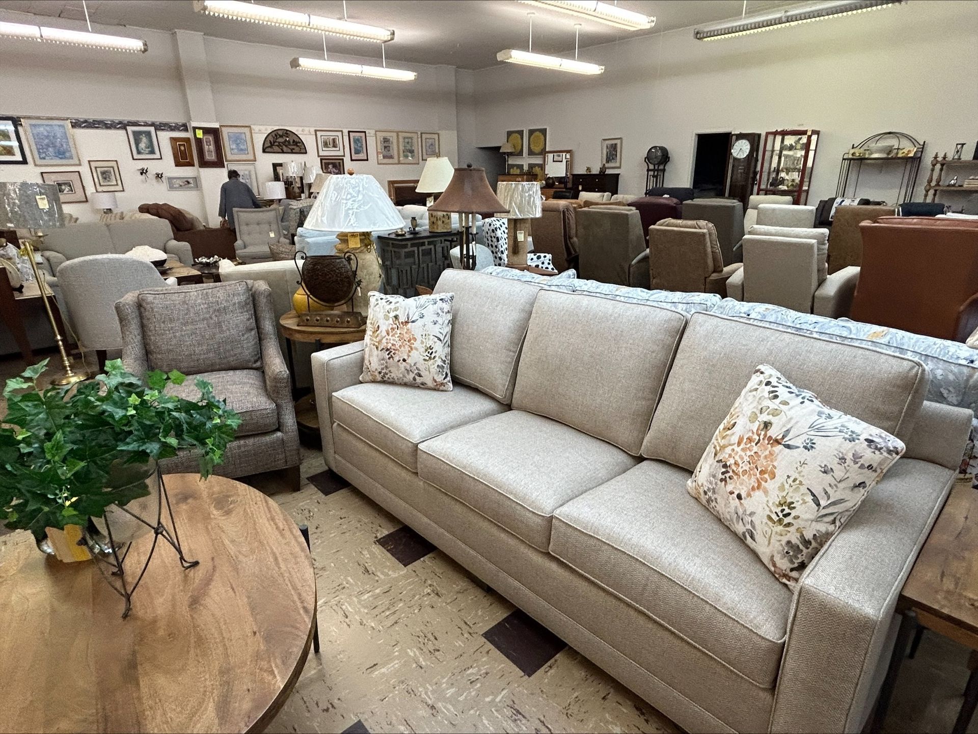 A furniture store showroom featuring a beige sofa, a patterned armchair, a round wooden table, and many display chairs.