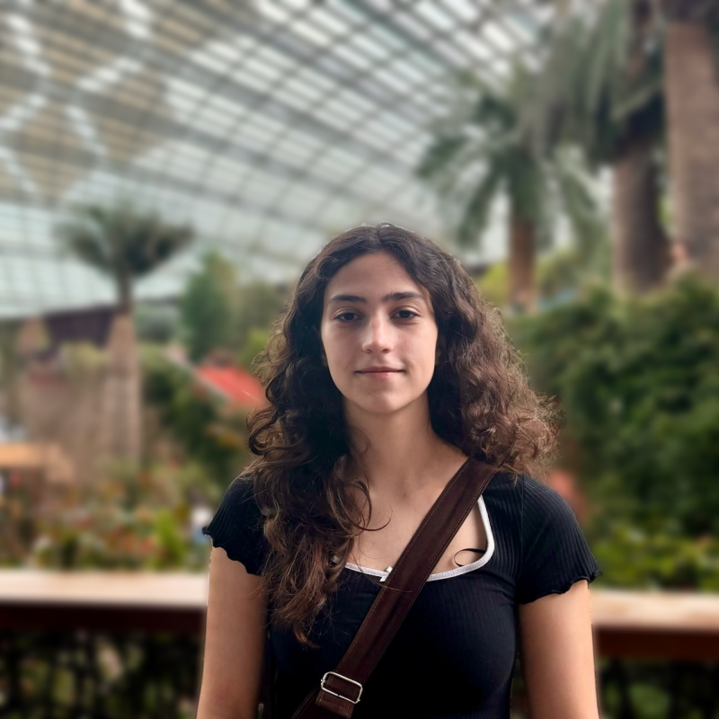 Young woman with brown curly hair, wearing a black top with a white trim, standing in a garden.