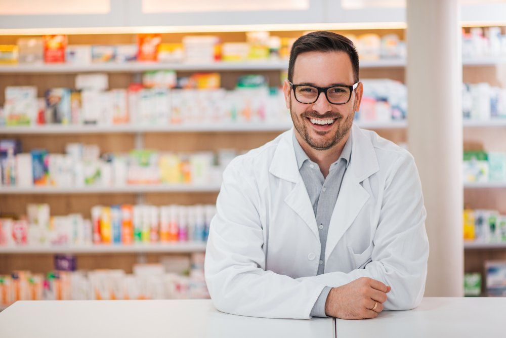 A smiling pharmacist is leaning on a counter in a pharmacy.