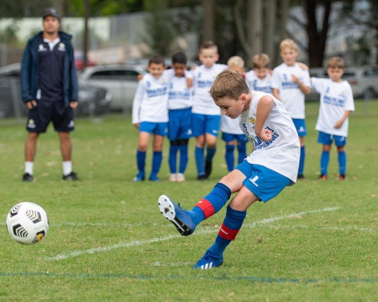 Young boy kicking a soccer ball while teammates watch during junior training