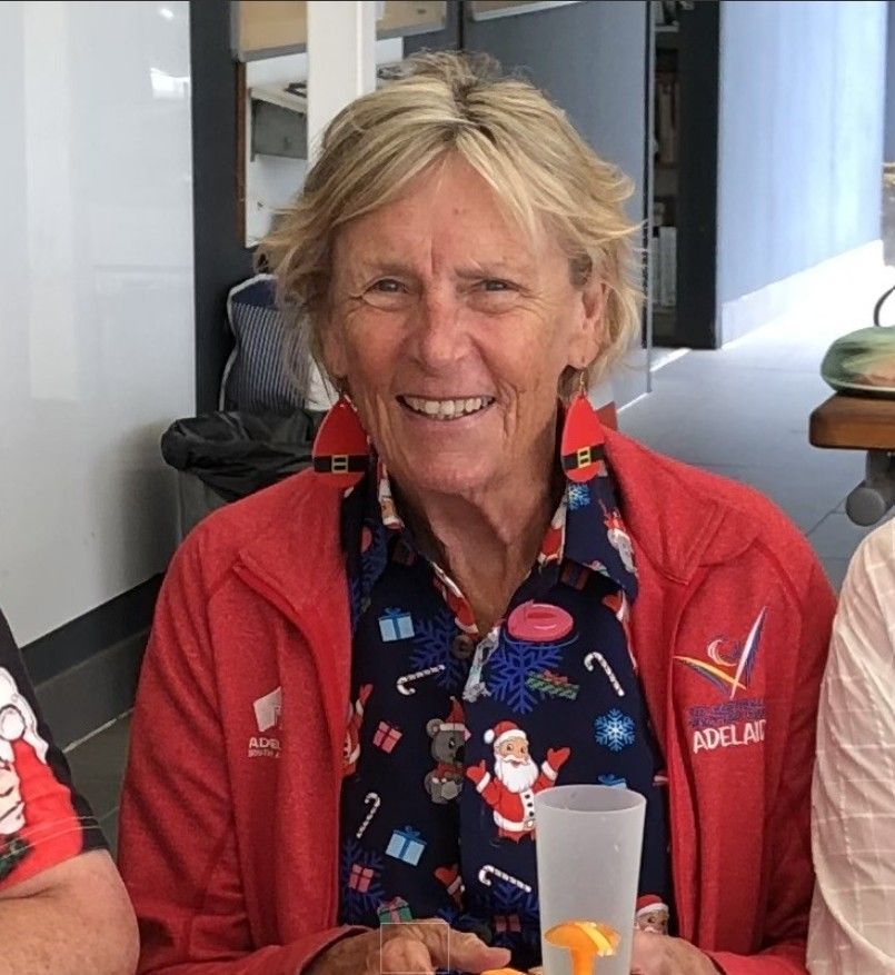 Sue Lewis smiling indoors while wearing a red jacket and patterned scarf