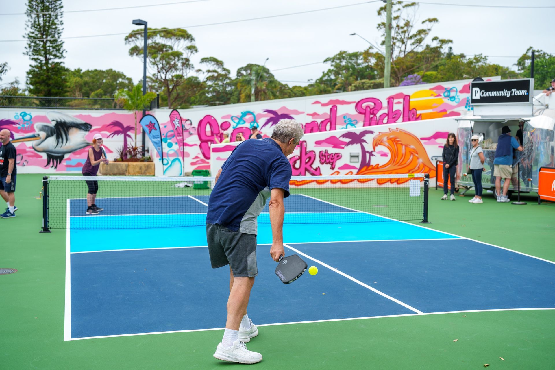 Player practicing a serve on a blue outdoor pickleball court with mural backdrop