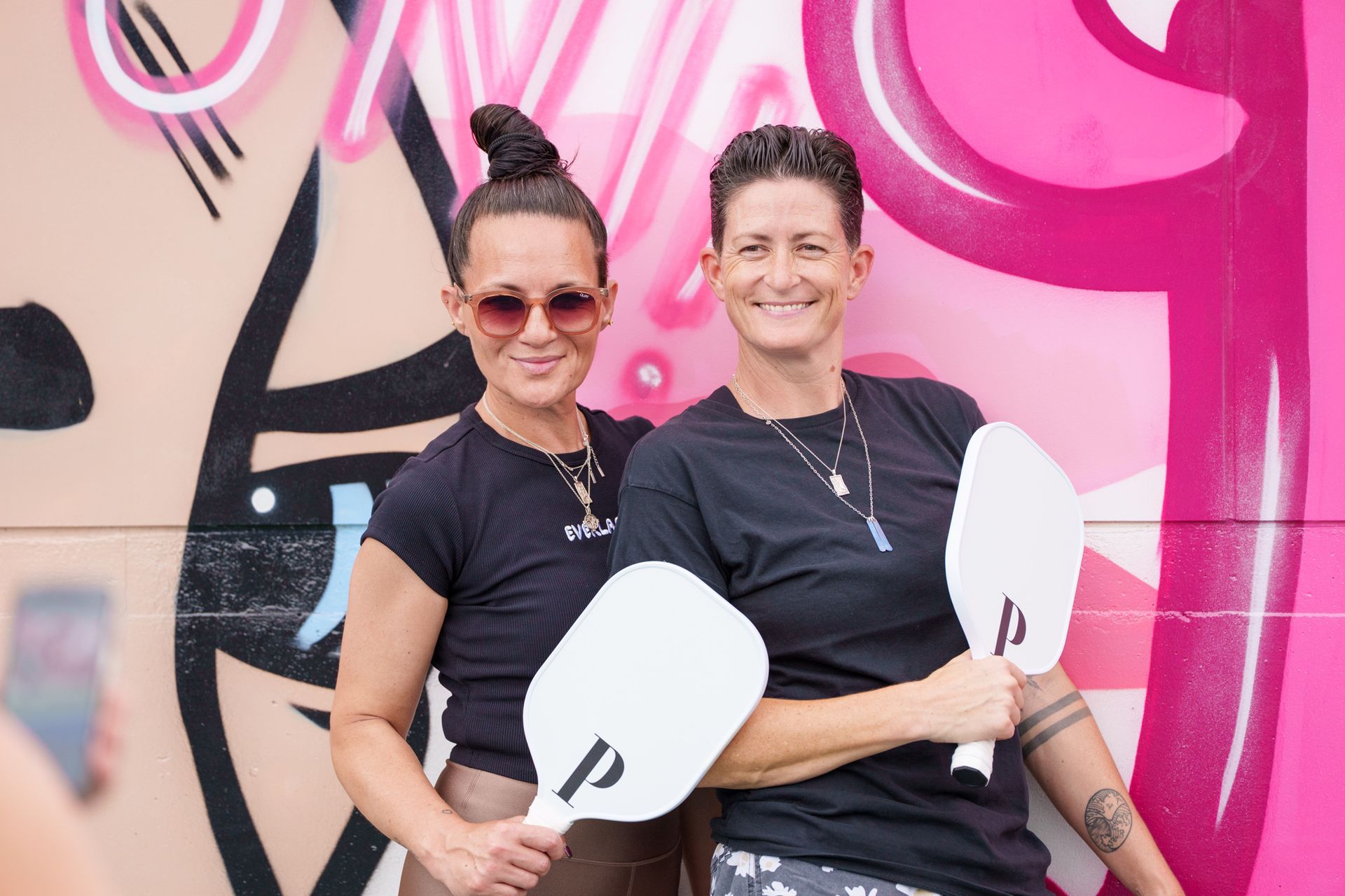 Two women smiling together while holding pickleball paddles beside vibrant court mural