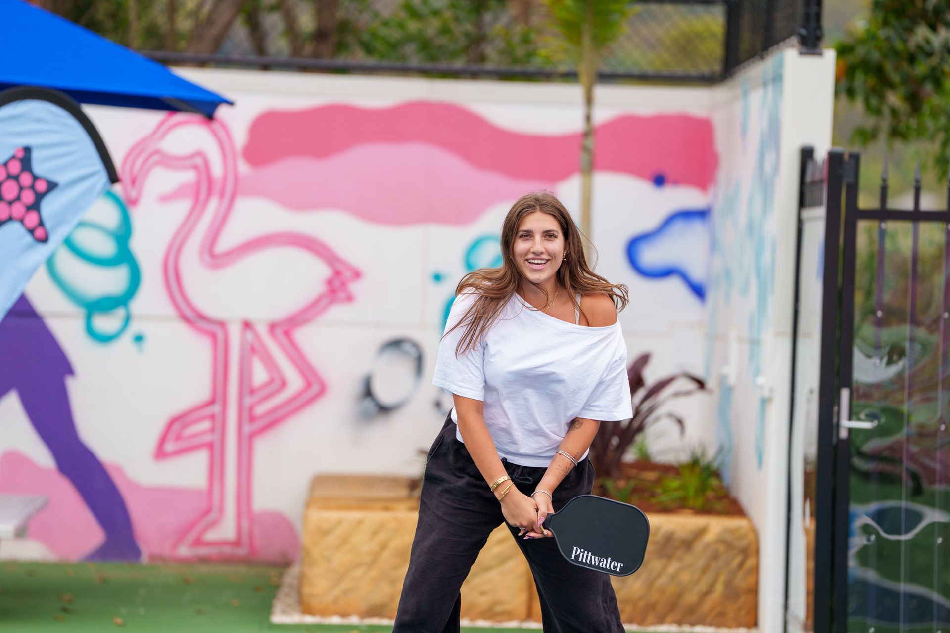 Smiling woman holding a pickleball paddle while standing near colourful court mural
